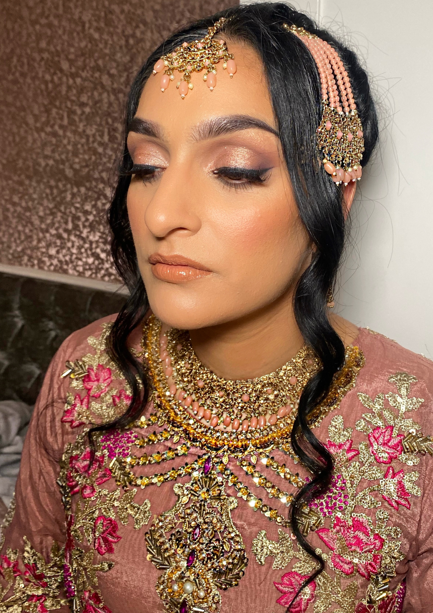 A woman dressed in pink traditional attire with elaborate gold and pink jewelry, including necklaces and a headpiece, has black wavy hair and makeup with soft eyeshadow and glossy lips.