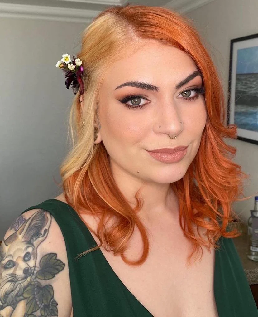 A woman with red hair styled in loose curls, wearing a dark green top, with flowers in her hair, visible tattoo on her shoulder, and a neutral makeup look.
