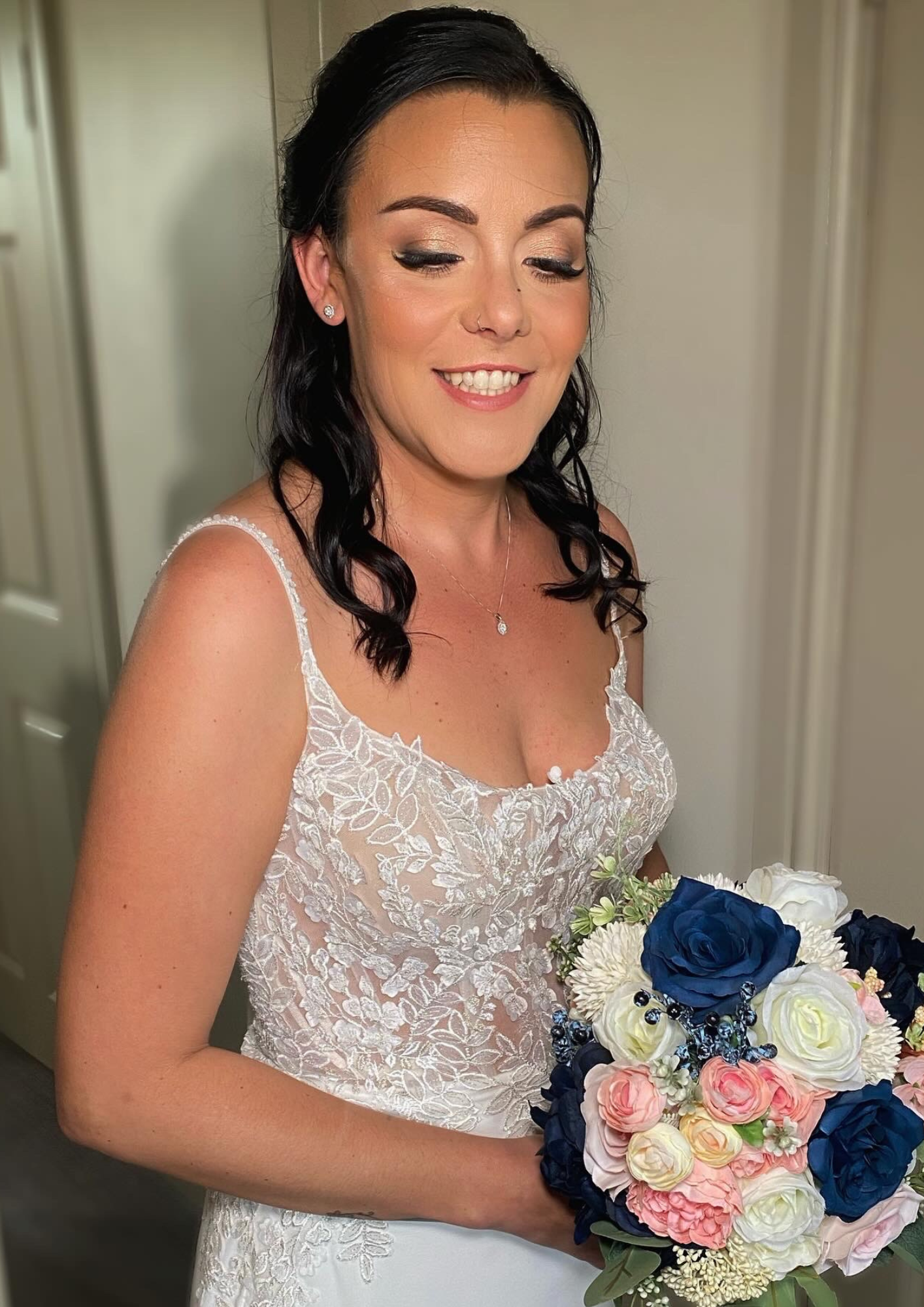 A smiling bride with dark wavy hair, wearing a lace wedding dress, holding a bouquet of white, pink, and navy blue roses.