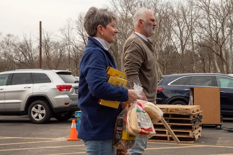Two people standing in a parking lot carrying bags of groceries, with parked cars and pallets in the background.