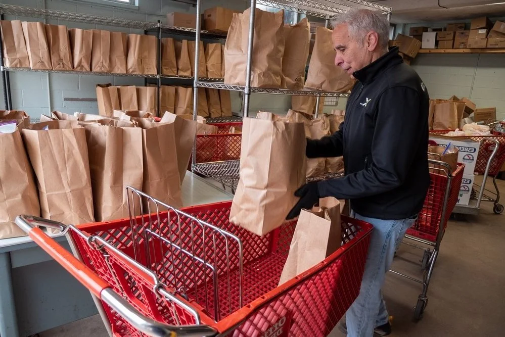 Man placing brown paper bags into a shopping cart in a storage area with shelves filled with similar bags.