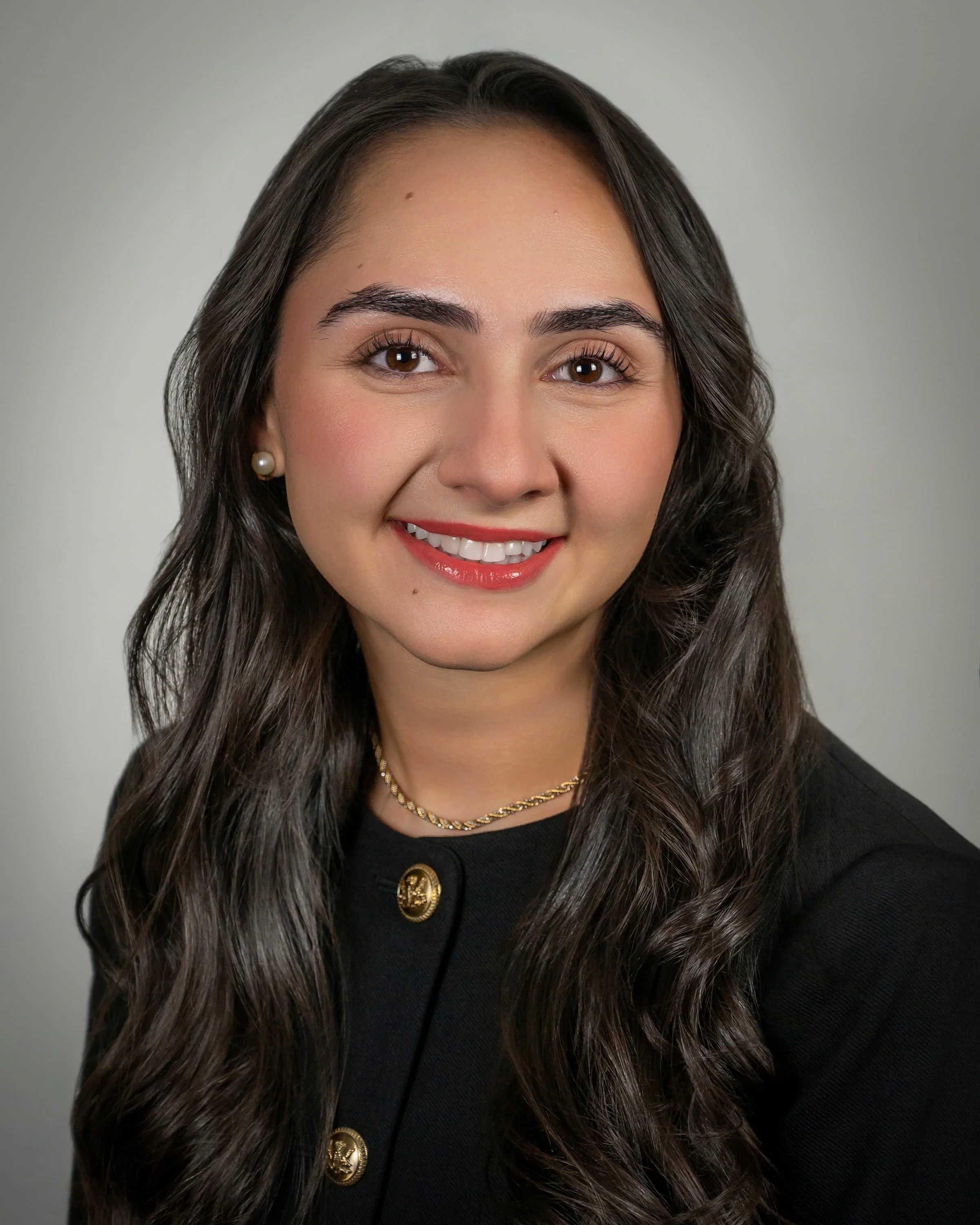 Portrait of a smiling woman with long, styled dark hair, wearing a black blazer, white blouse, and gold necklace, against a light gray background.
