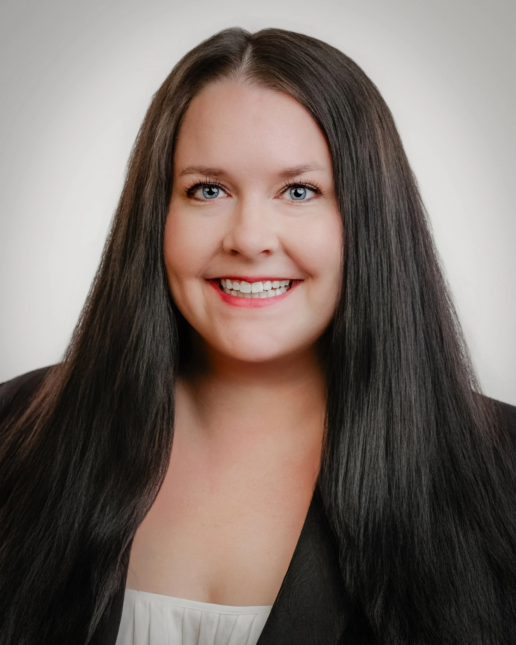 Close-up portrait of a woman with long dark hair, blue eyes, and a friendly smile, wearing a black top against a plain light background.