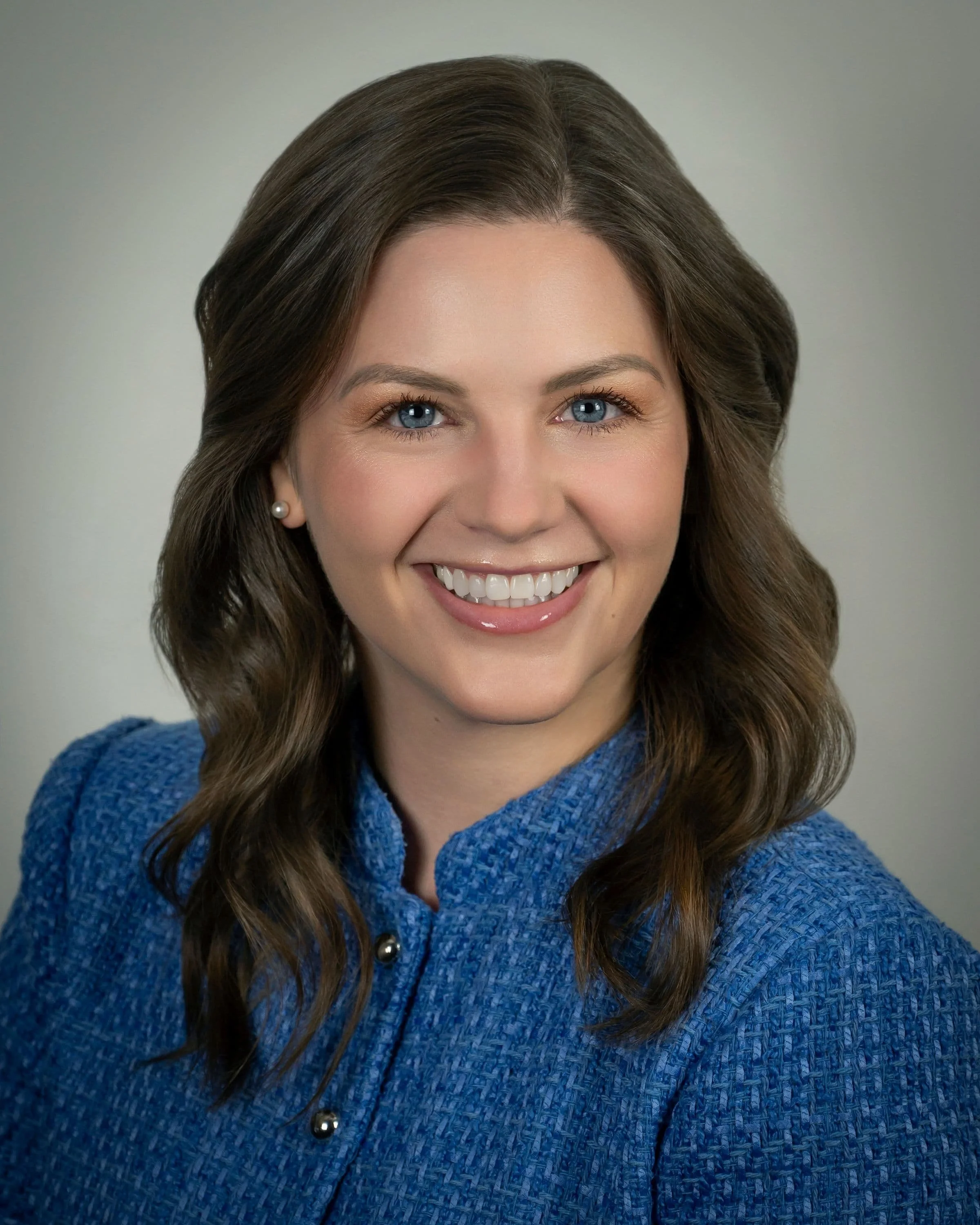 Headshot of a woman with wavy brown hair, blue eyes, smiling, wearing a textured dark blazer with a gold brooch and pearl earrings, against a plain light gray background.