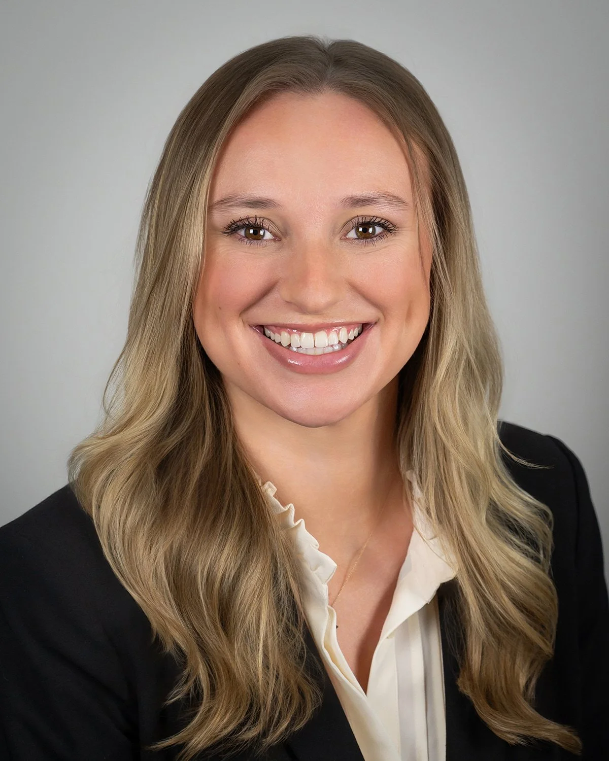 Headshot of a woman with long blonde hair, smiling, wearing a black blazer, light pink blouse, and a gold necklace with a small pendant.