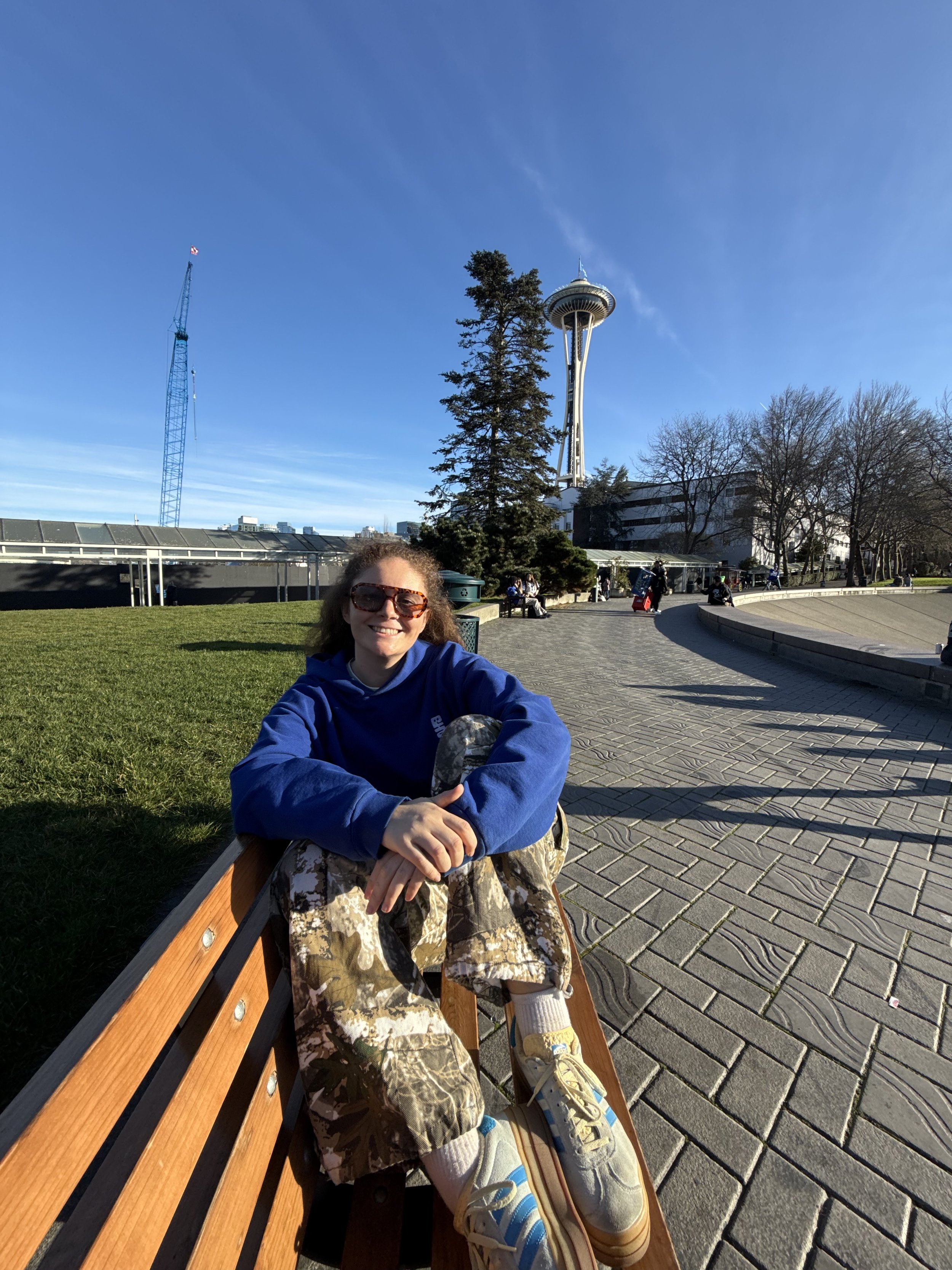 A woman sitting on a park bench on a sunny day, smiling at the camera, with the Space Needle in the background in Seattle, Washington.