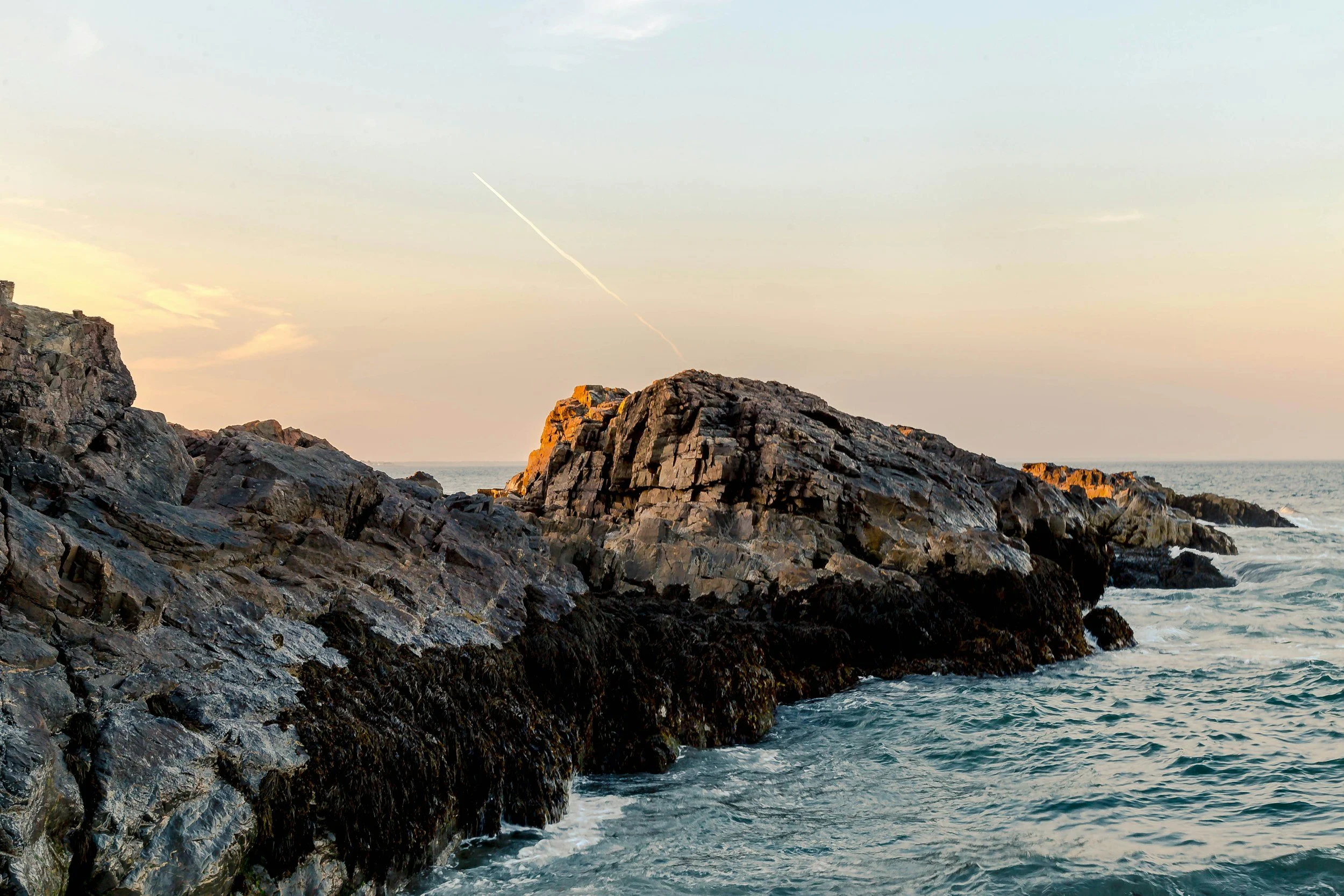 Rocky coastline with waves crashing against dark, jagged rocks during sunset, with a light blue sky and some clouds.