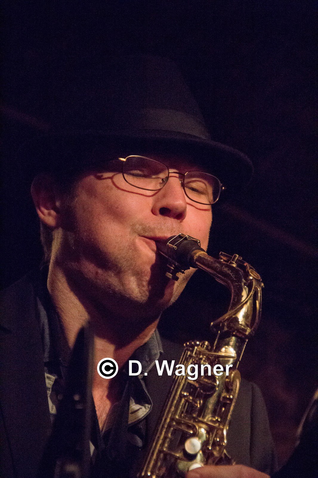 A man wearing glasses and a hat playing a saxophone in a dimly lit setting.