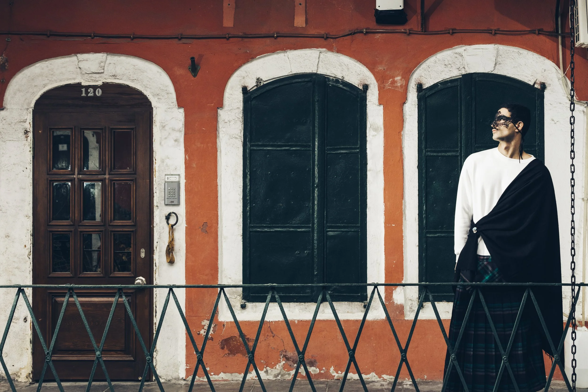 Person standing outside an old building with red walls and arched windows, wearing a white shirt, plaid kilt, black shawl, and decorative mask.