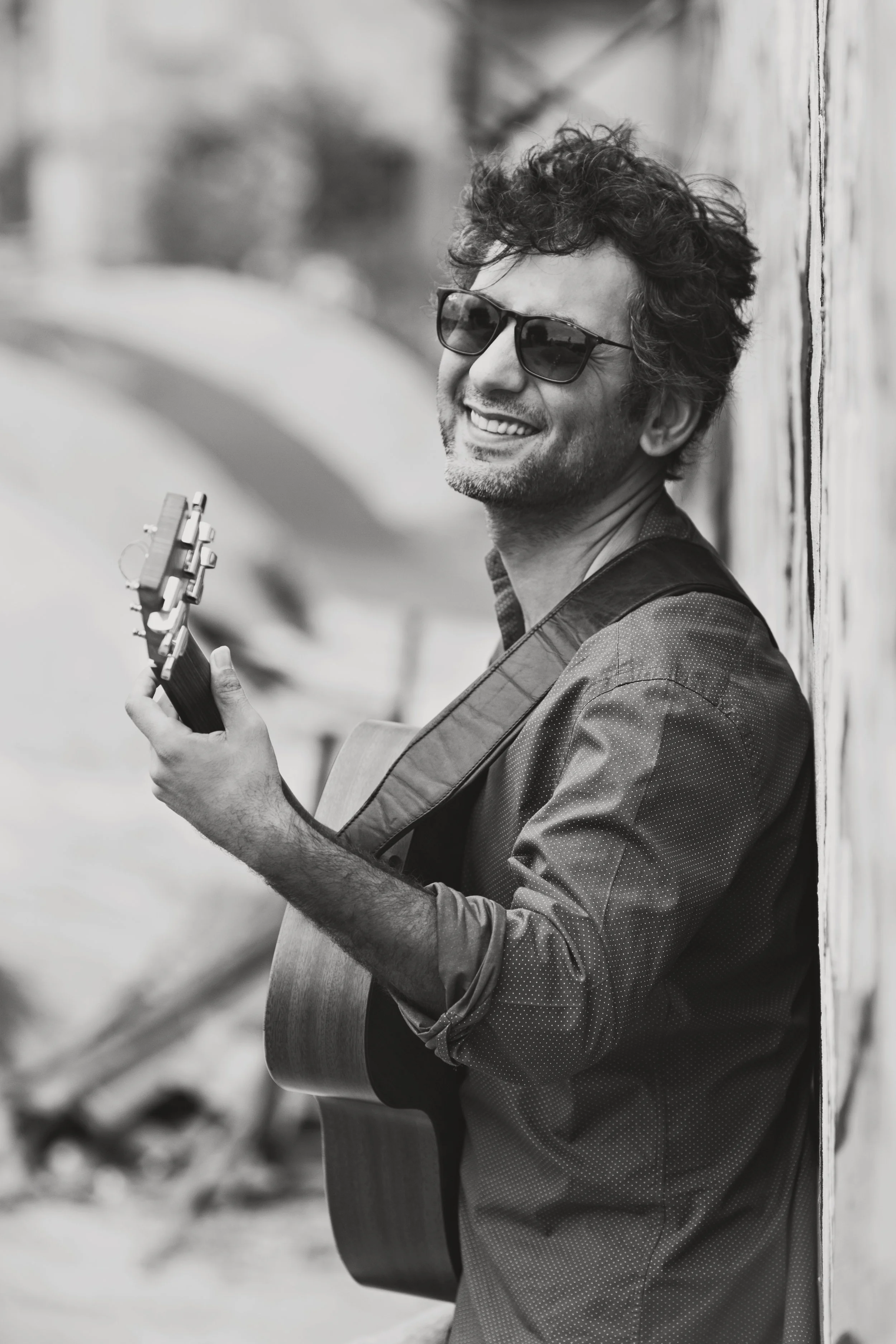 Black and white photo of a man playing guitar outdoors, wearing sunglasses and smiling.