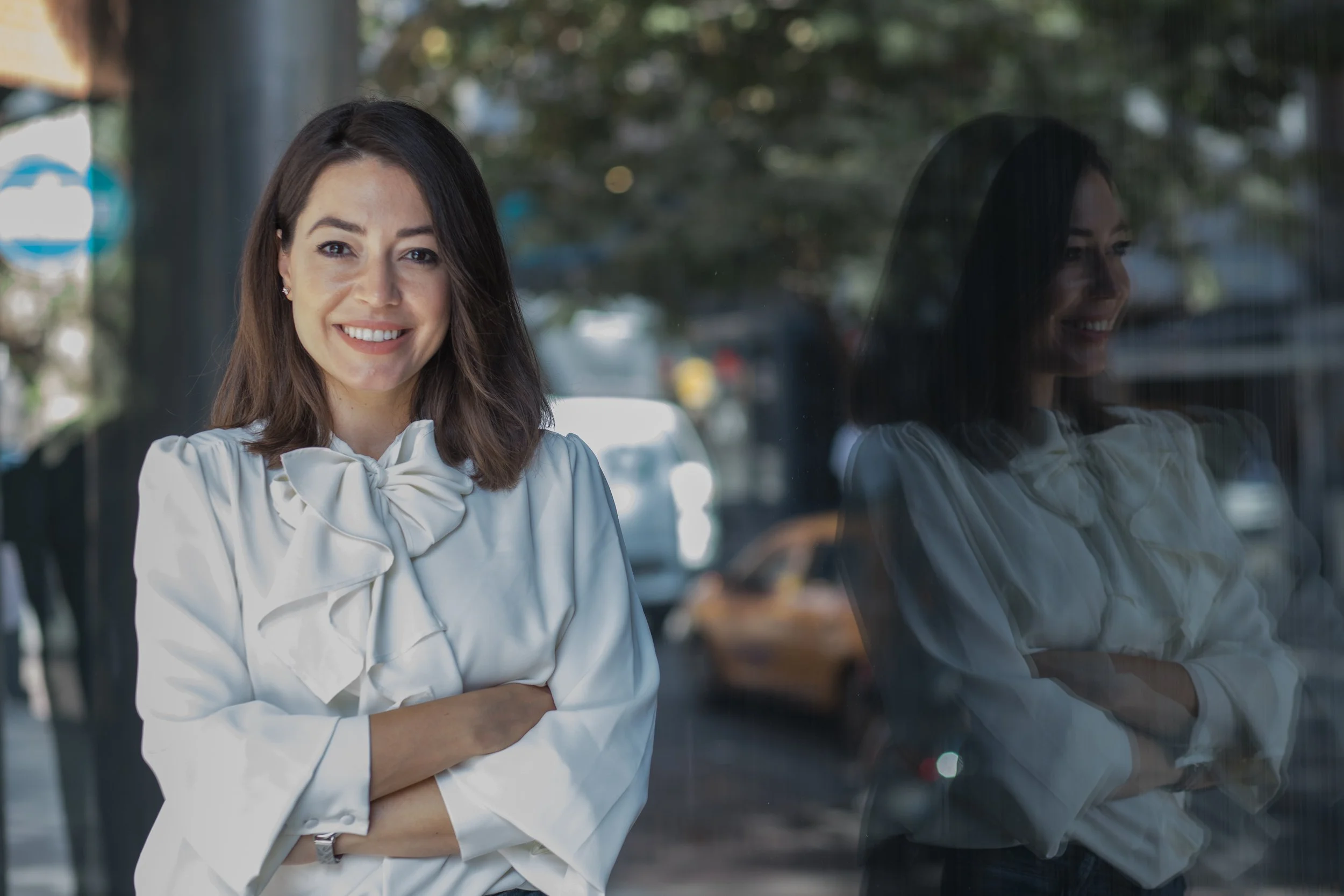 Woman in white blouse smiling with arms crossed, standing by a window showing her reflection. Background includes city street with trees and parked cars.