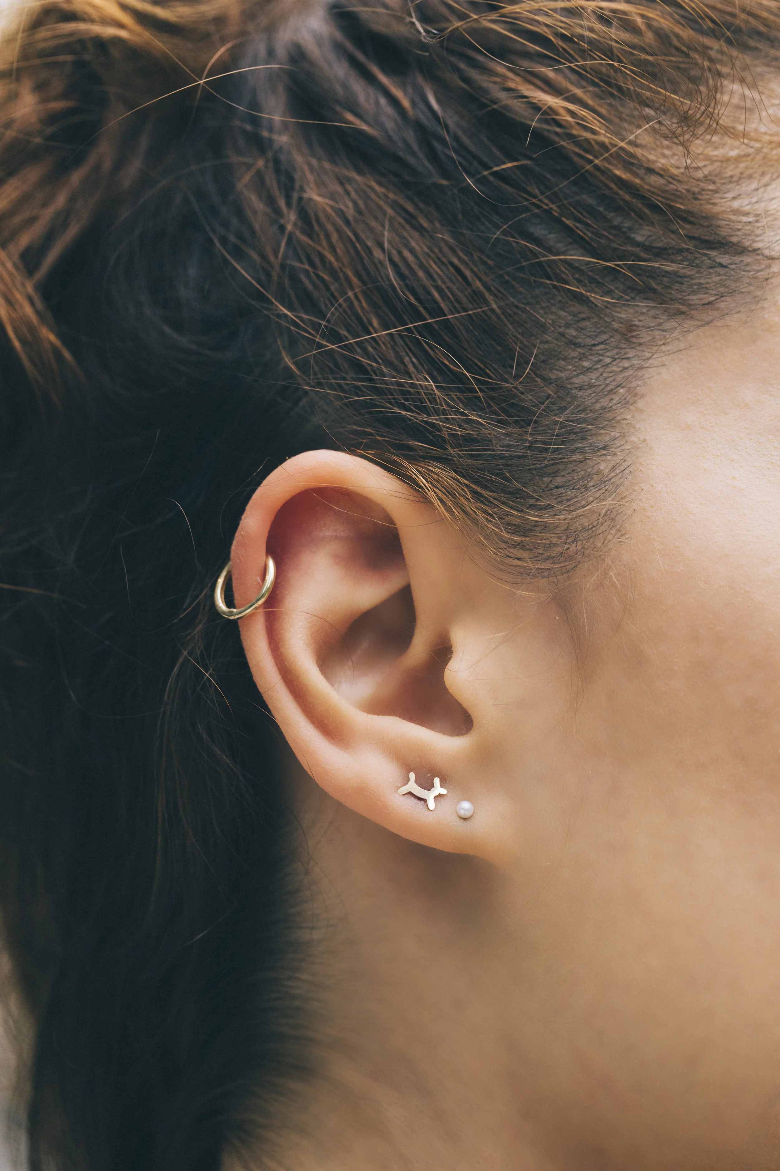 Close-up of an ear with three earrings: a hoop in the cartilage, a small abstract design in the lobe, and a pearl stud below it.