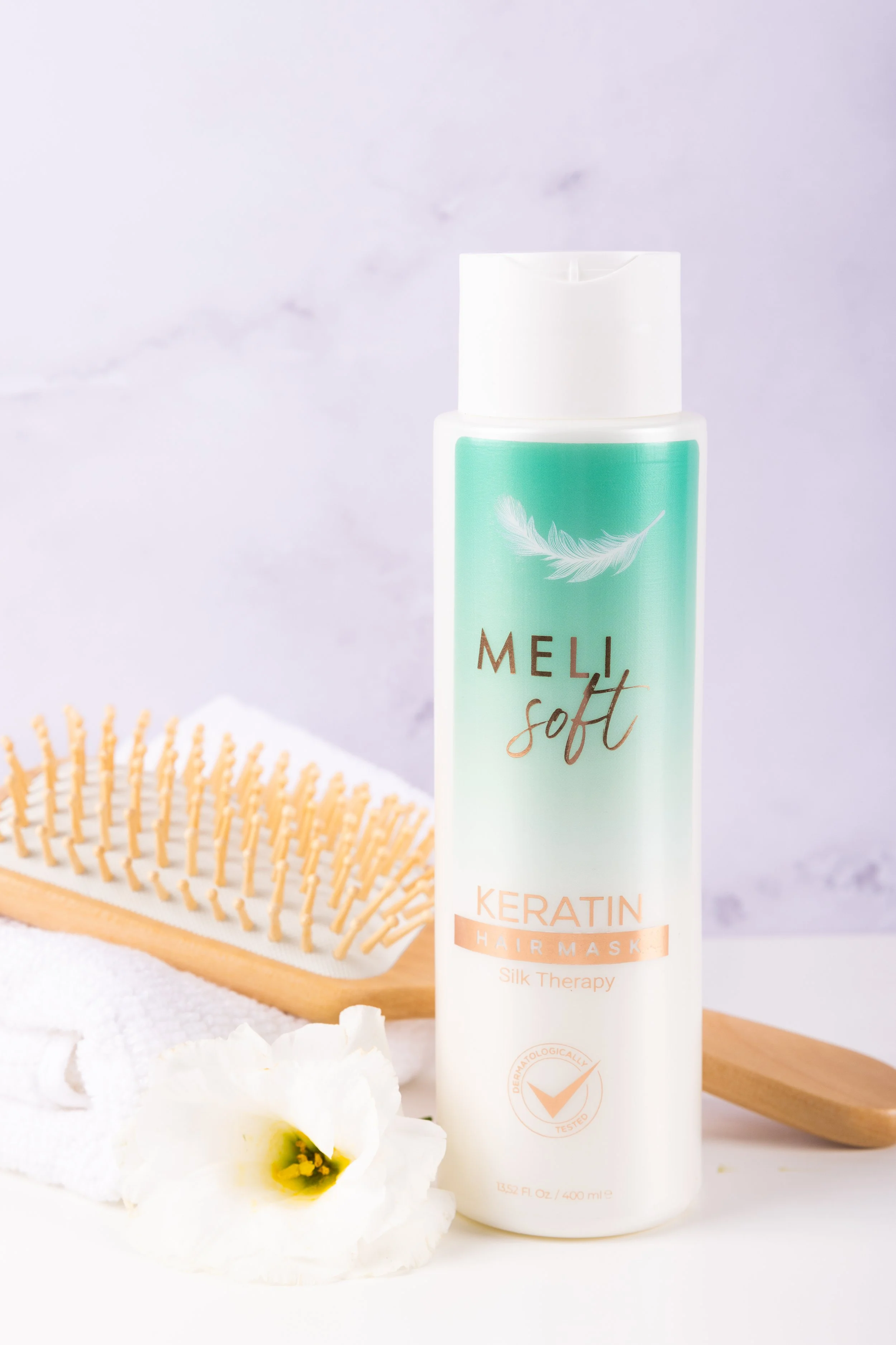Bottle of Meli Soft Keratin Hair Mask and a wooden hairbrush on a white towel with a white flower.