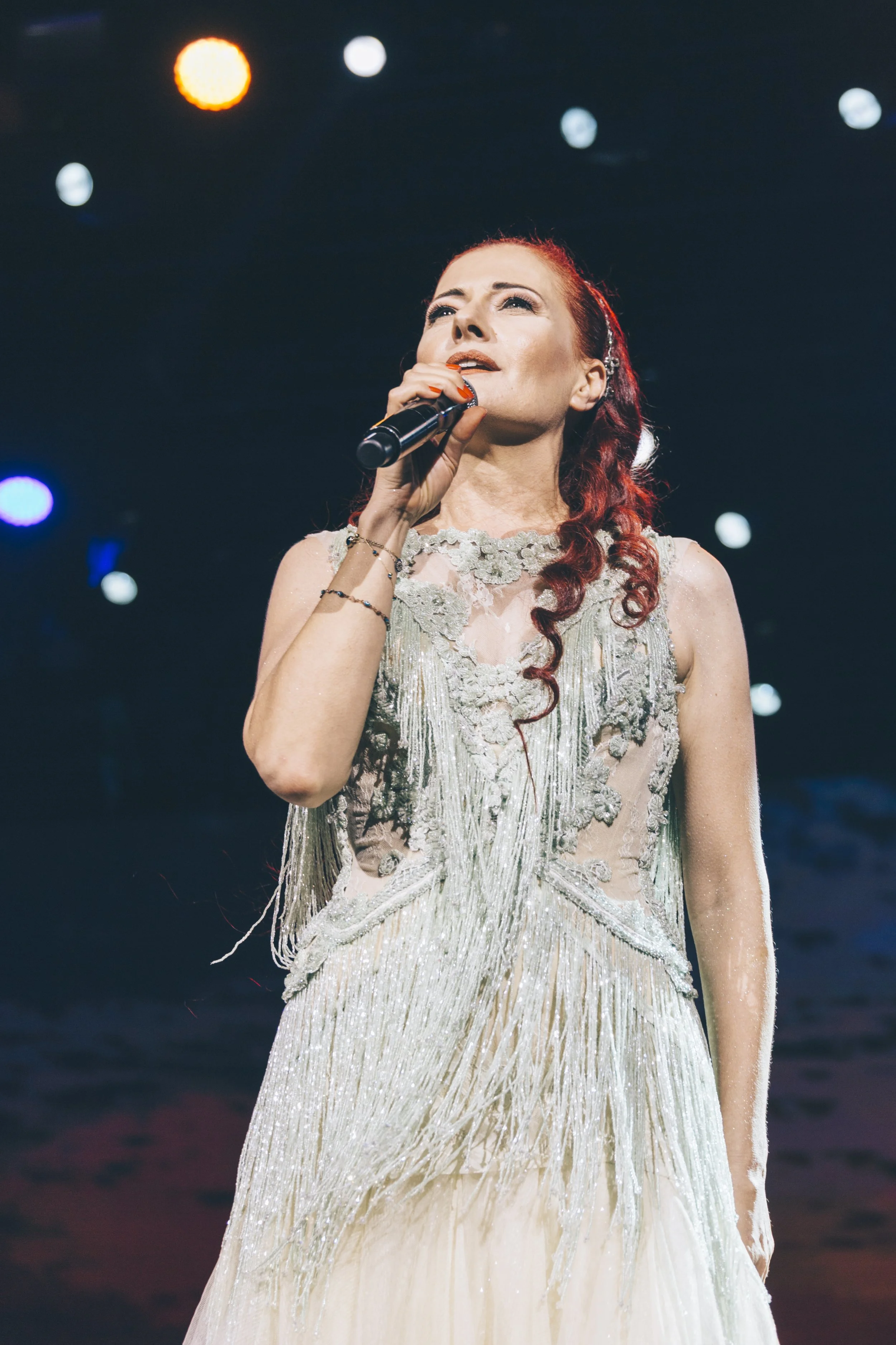 Singer performing on stage in an elegant dress with microphone, vibrant lights in background.