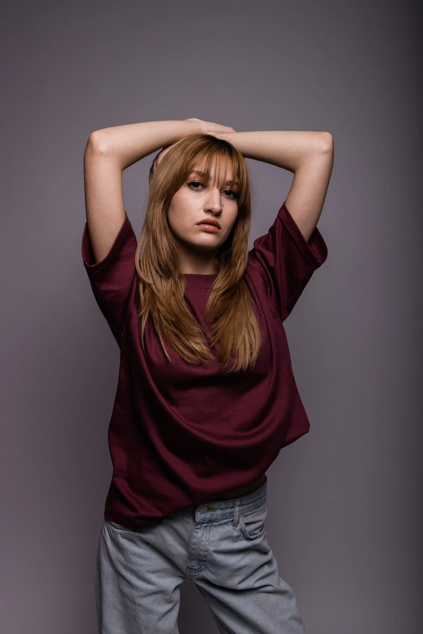 Person with long hair wearing a maroon shirt and blue jeans, posing with arms above their head against a gray background.