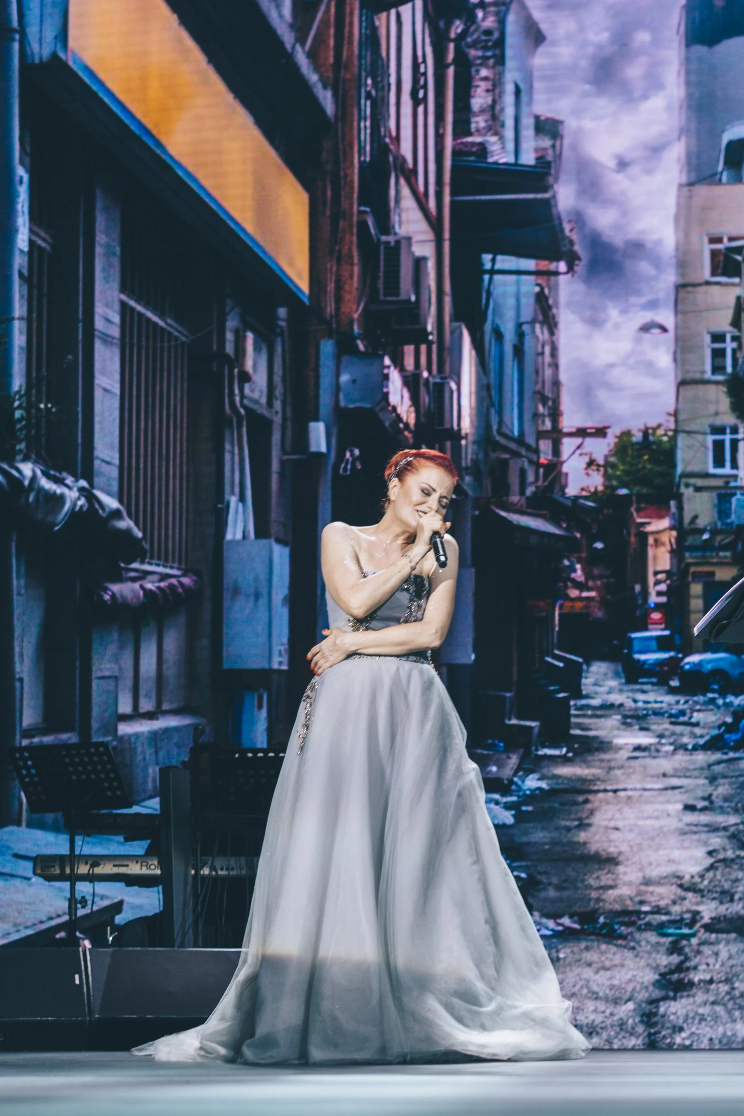 Singer with red hair performing on stage, wearing a gray gown, urban background