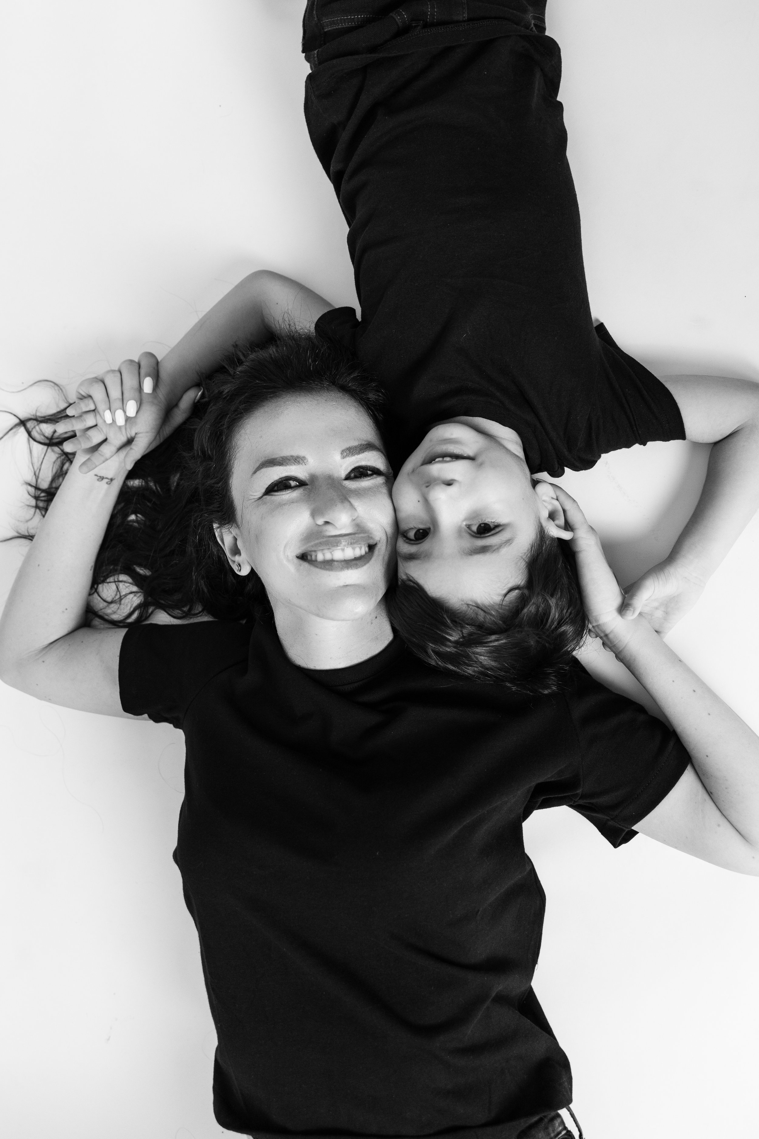 A black and white photo of a woman and a child lying on the floor, facing each other, both smiling. They are wearing black shirts, and their hands are gently touching each other's heads.