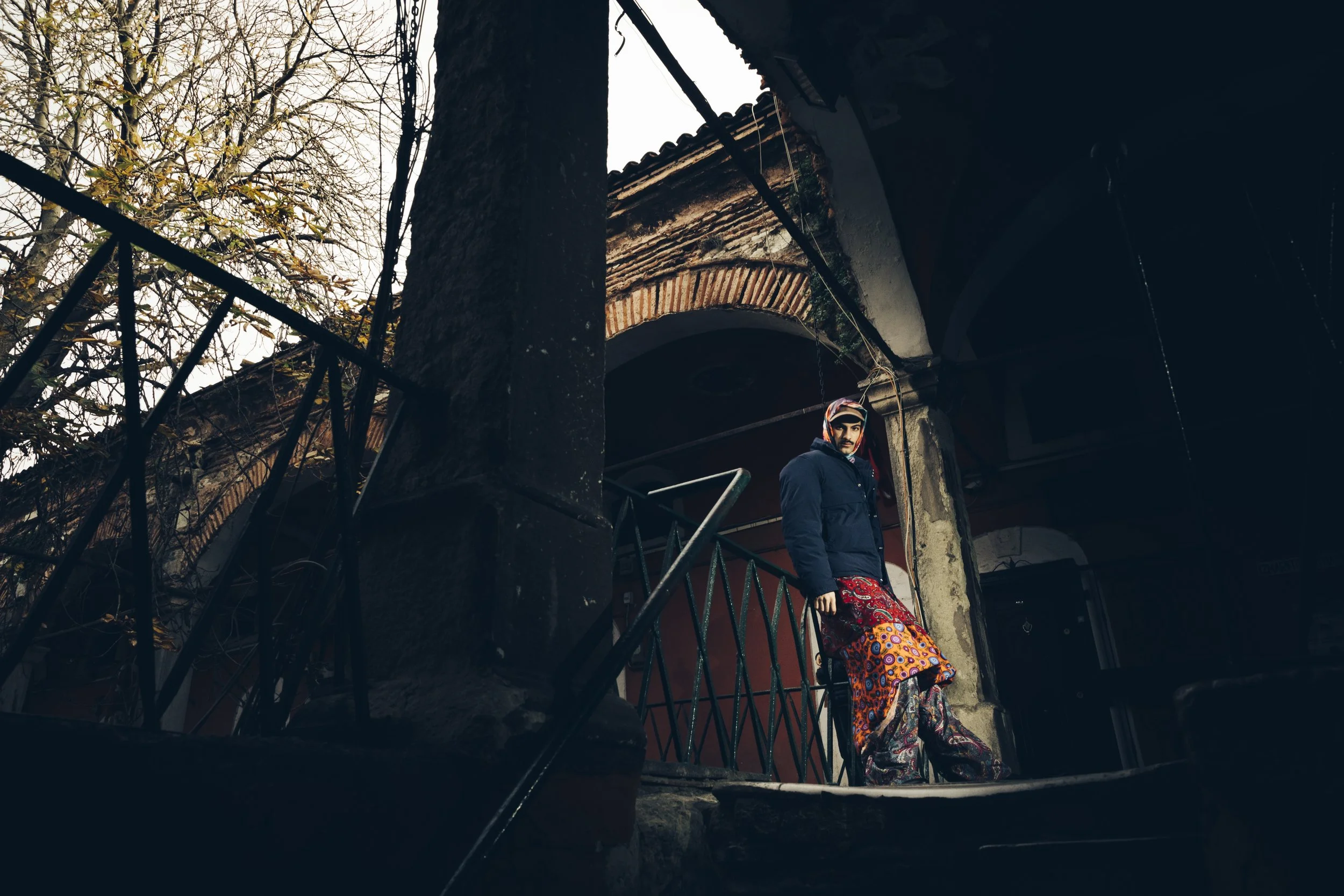 Person in colorful attire standing on steps in a dimly lit, old architectural setting with a bare tree in the background.