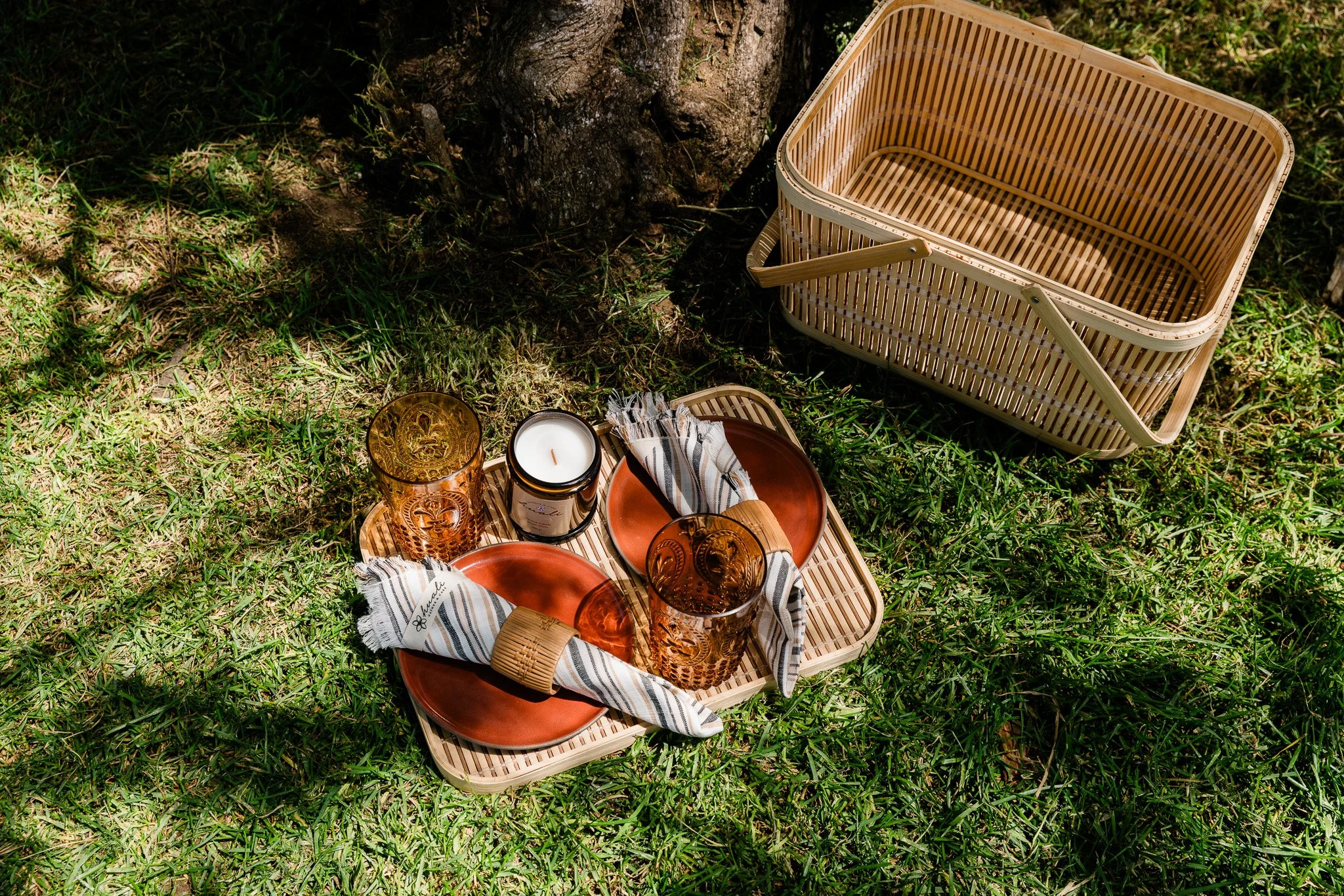 Outdoor picnic setup on grass with a wicker tray holding orange plates, amber glasses, striped napkins, and a candle; empty wicker basket beside.