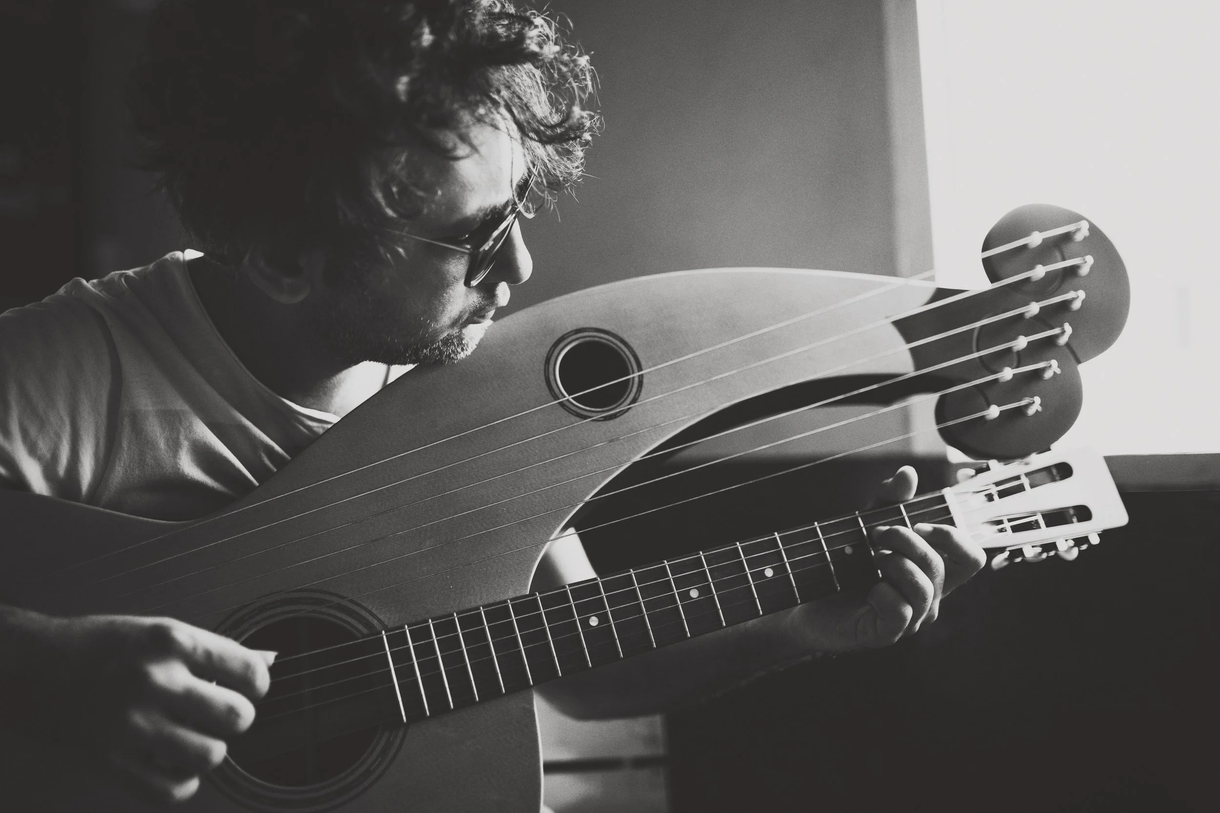 Black and white photo of a person playing a harp guitar in a dimly lit room, wearing sunglasses and focusing on the instrument.