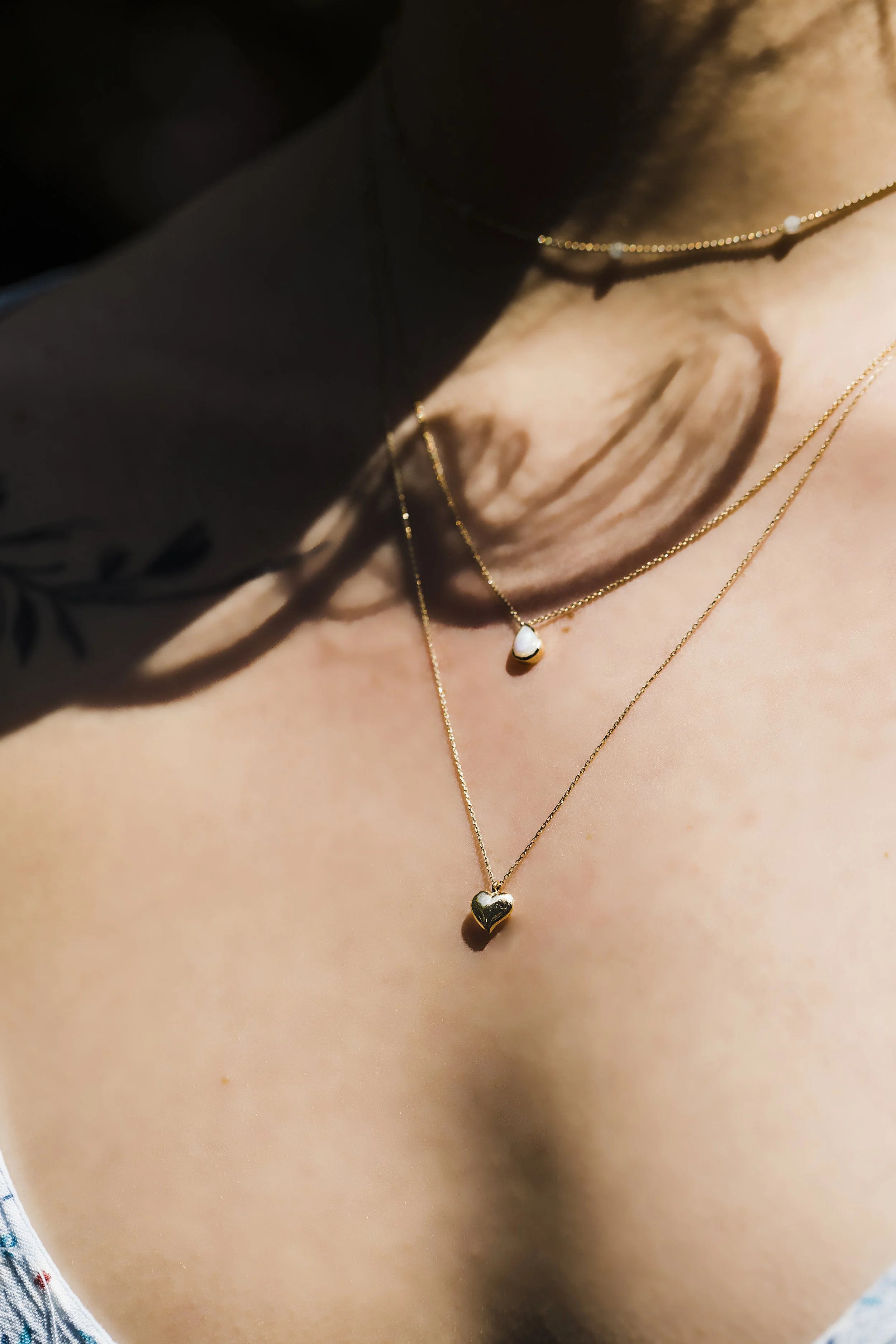 Close-up of a person's neck and upper chest wearing two gold necklaces, each with a small heart pendant, with natural lighting and shadows.