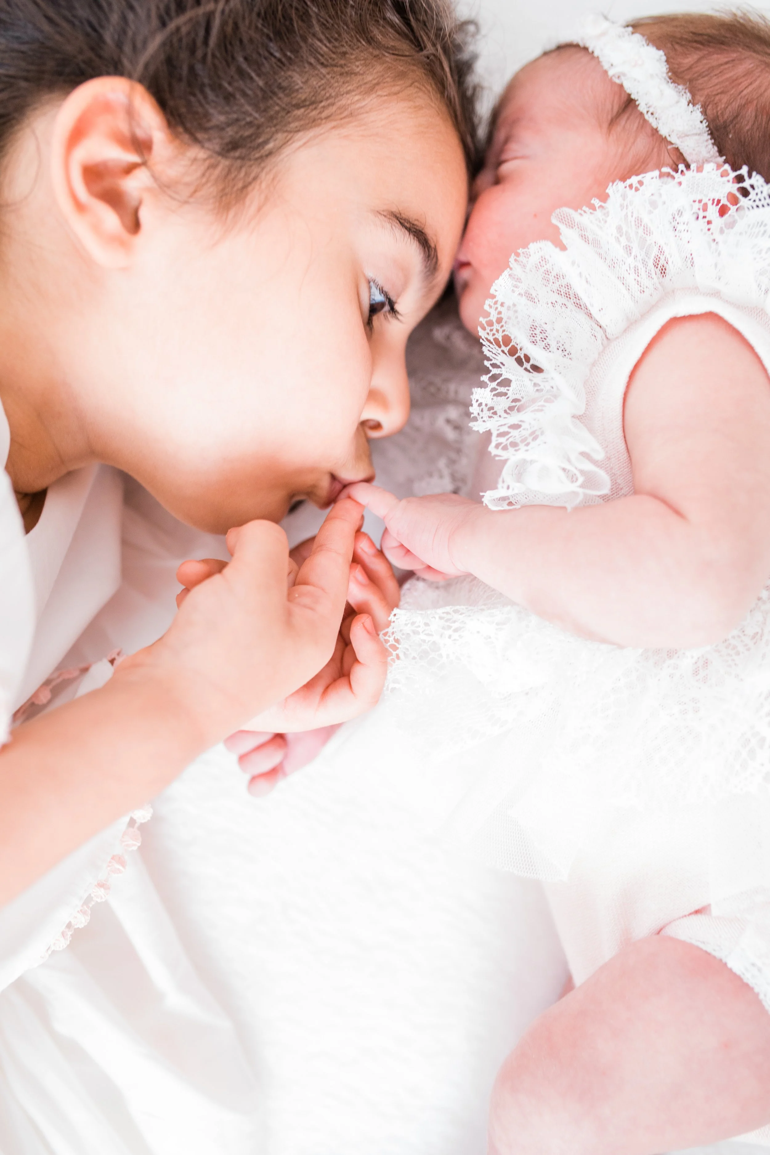Young child gently holding and kissing a newborn baby's hand, both dressed in white lace garments, on a soft background.