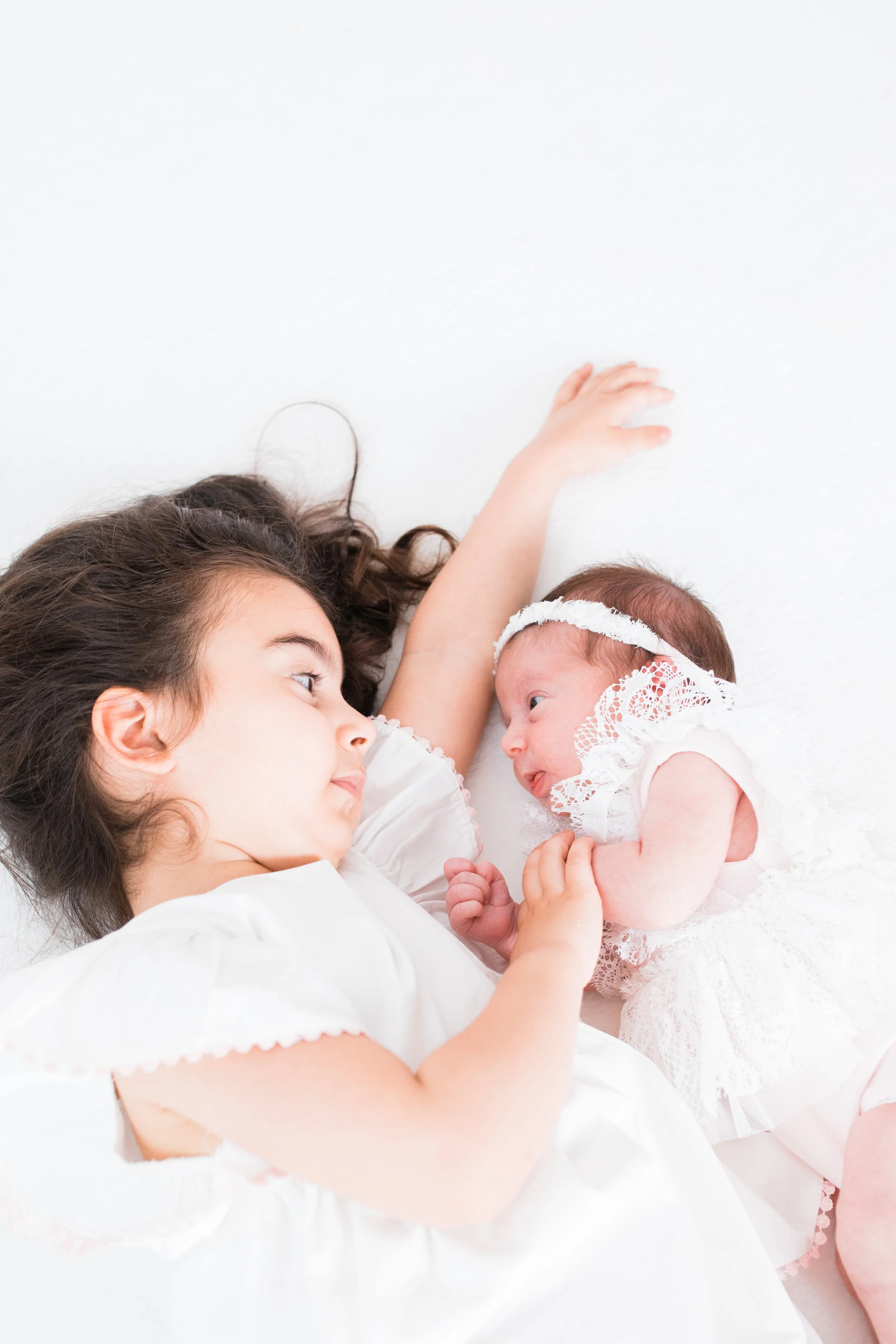 Young girl lying next to a newborn baby wearing white lace outfit.