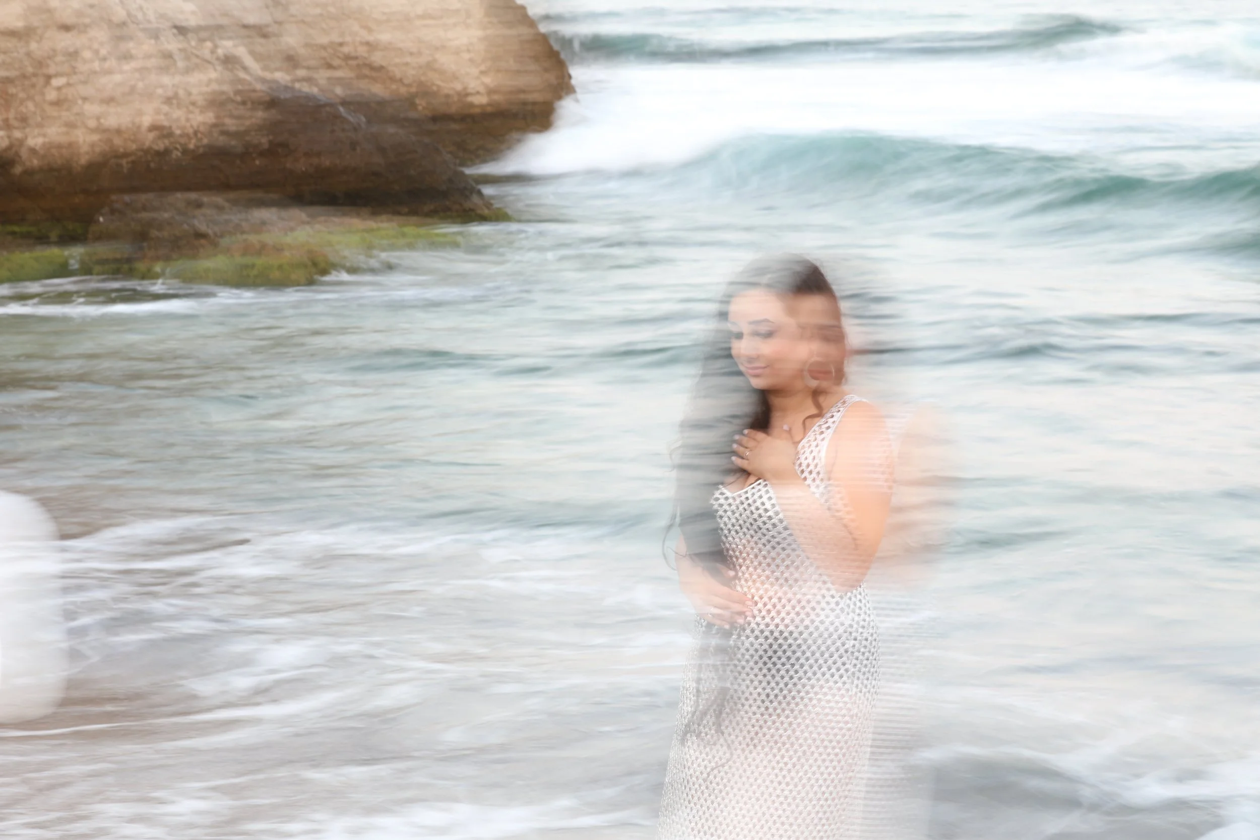 Blurred image of a woman standing by the seashore with rocky outcrop in the background.