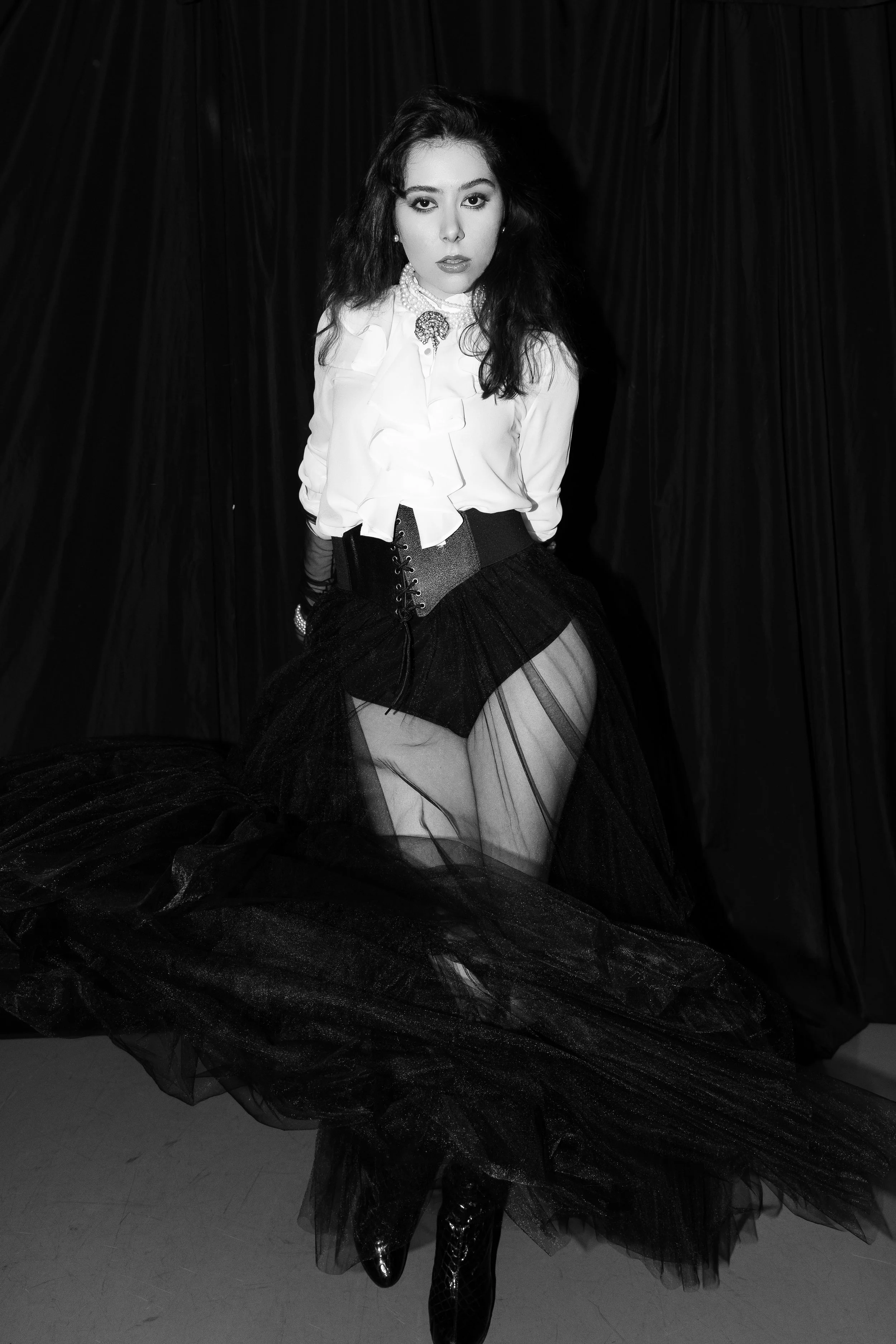 Person wearing a white blouse and black sheer skirt, standing against a dark curtain backdrop in a black and white photo.