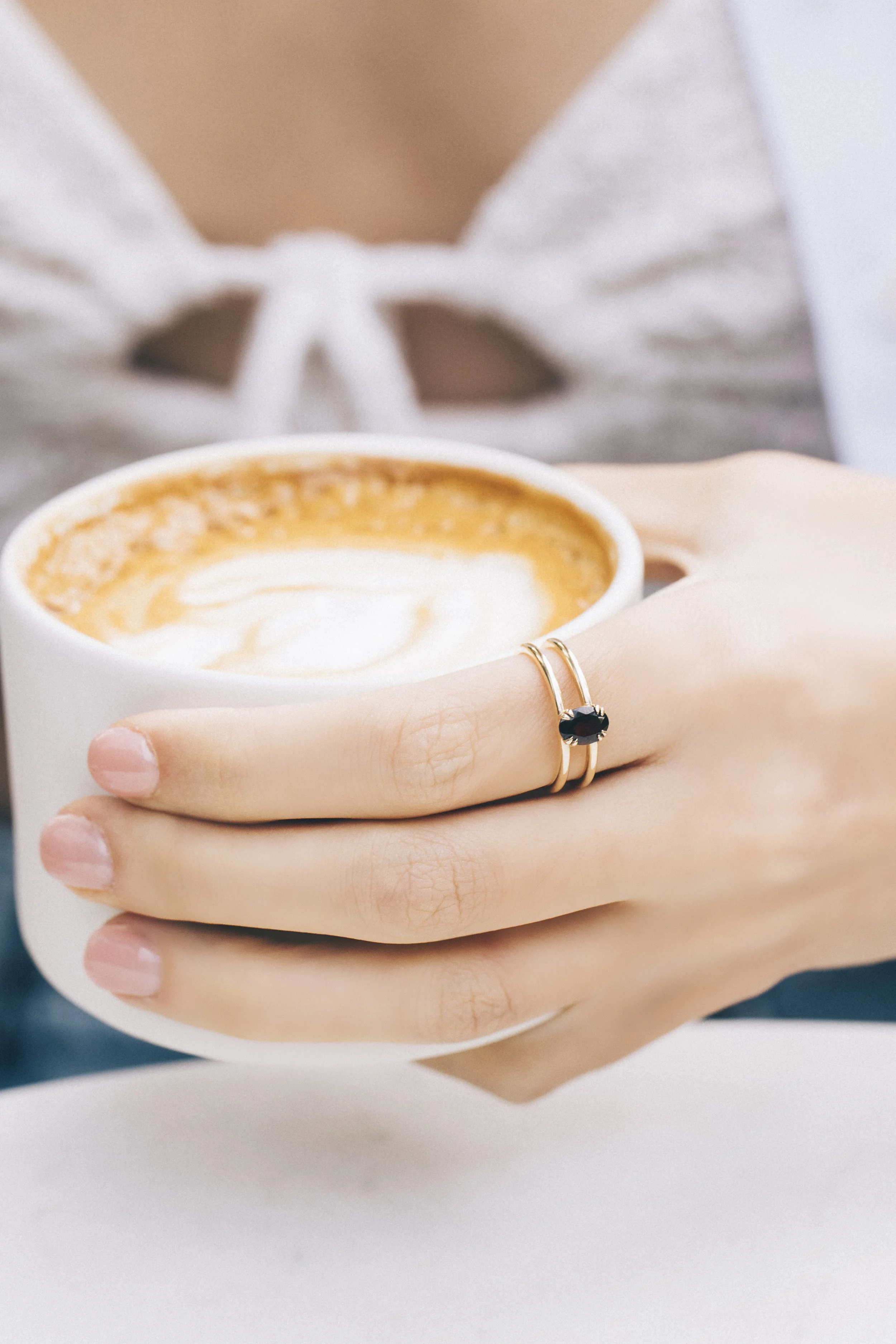Close-up of a person's hand with gold rings holding a cup of coffee with latte art.