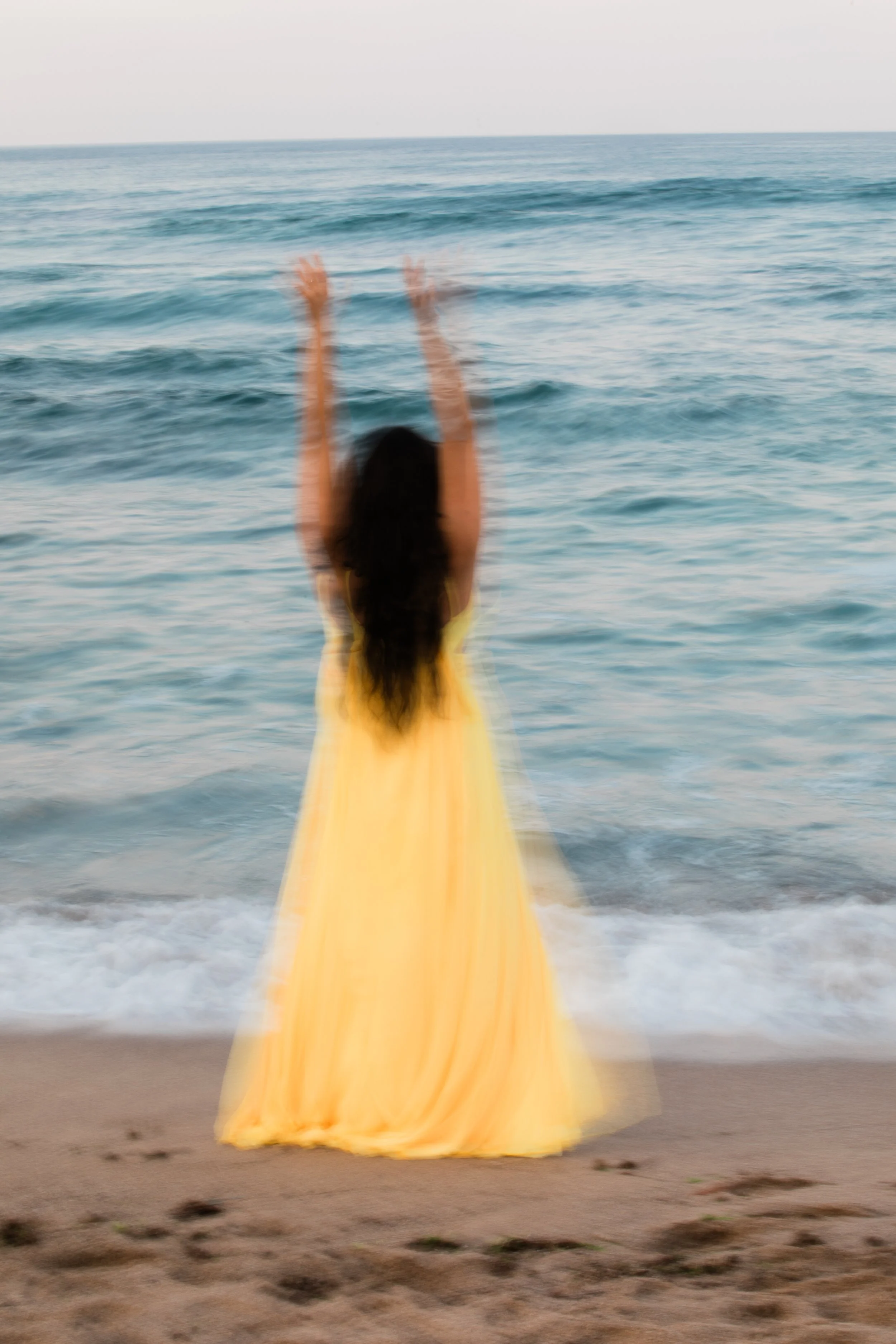 Blurred image of a person in a yellow dress raising their arms on a beach with waves in the background.