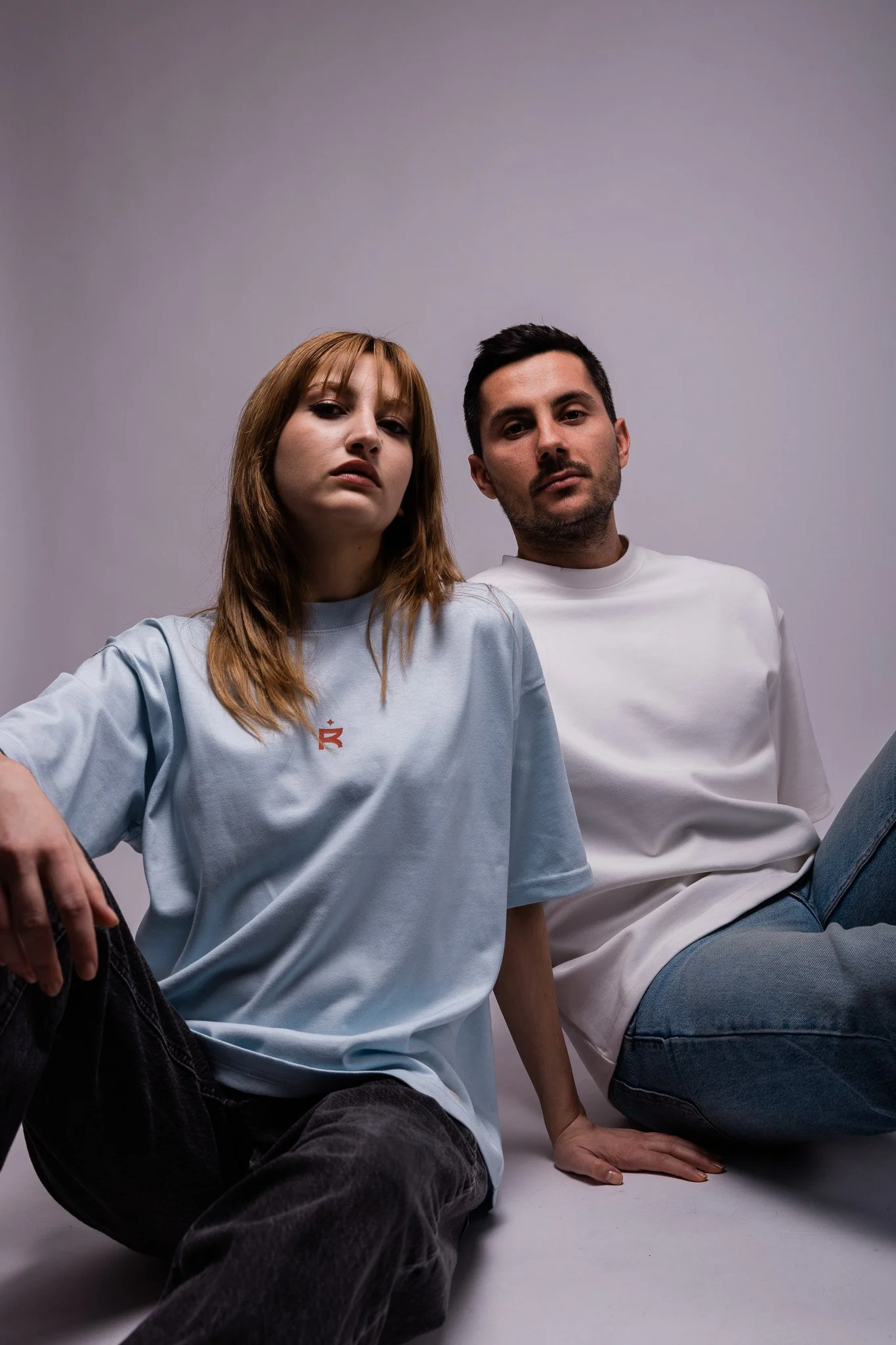 Two people sitting, wearing casual t-shirts and jeans, with a neutral background.