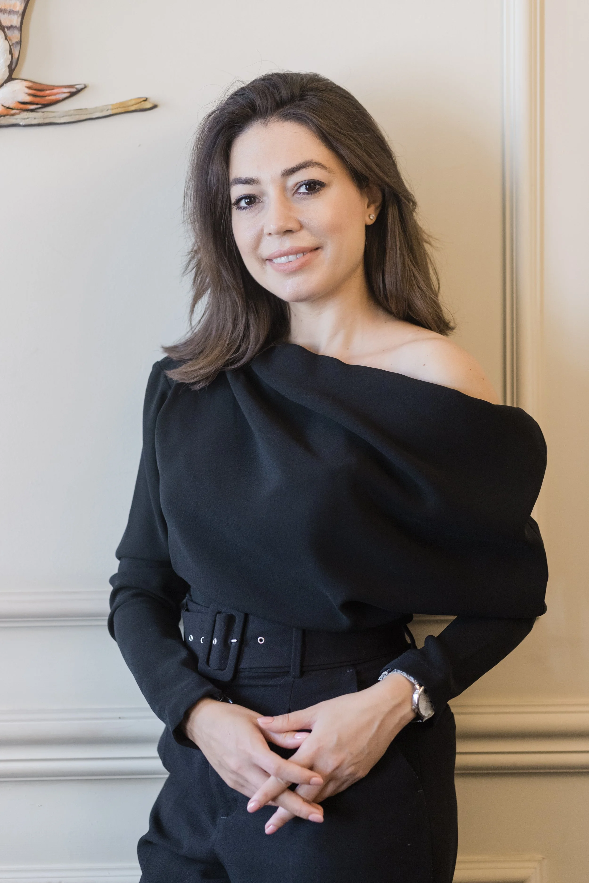 A woman wearing a black off-the-shoulder top and belted trousers, standing against a light-colored wall with decorative molding and artwork partially visible, posing with hands clasped.