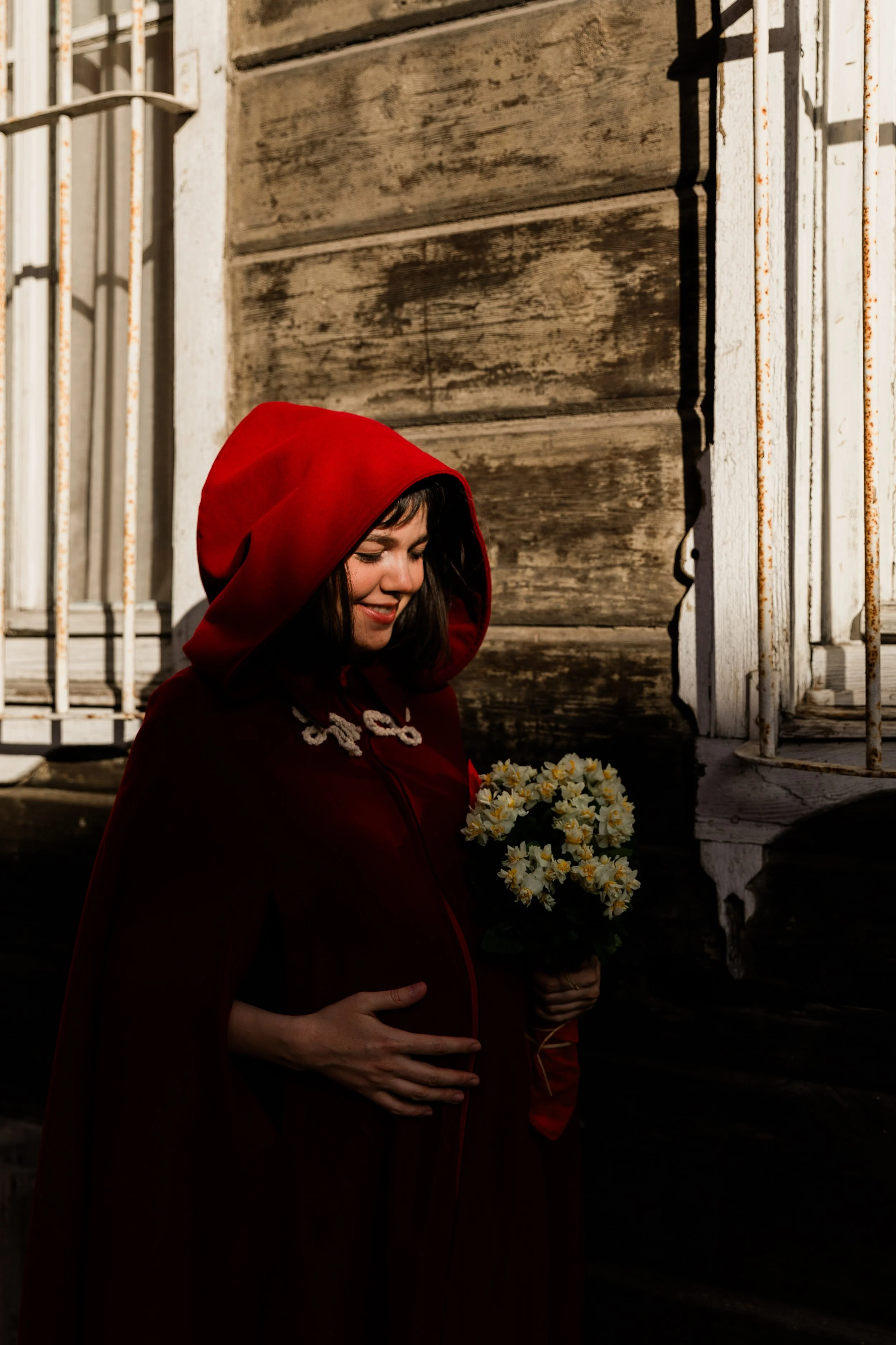Person wearing a red hooded cloak holding a bouquet of white flowers against a rustic wall.