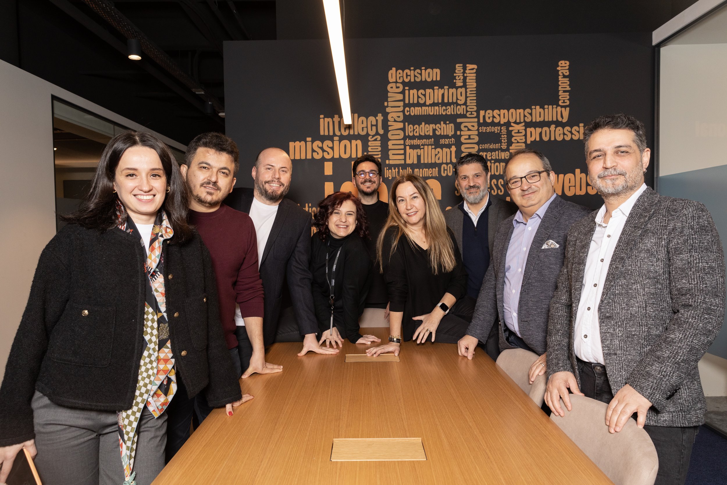 Group of people smiling and posing in an office setting with a conference table and wall of motivational words in the background.