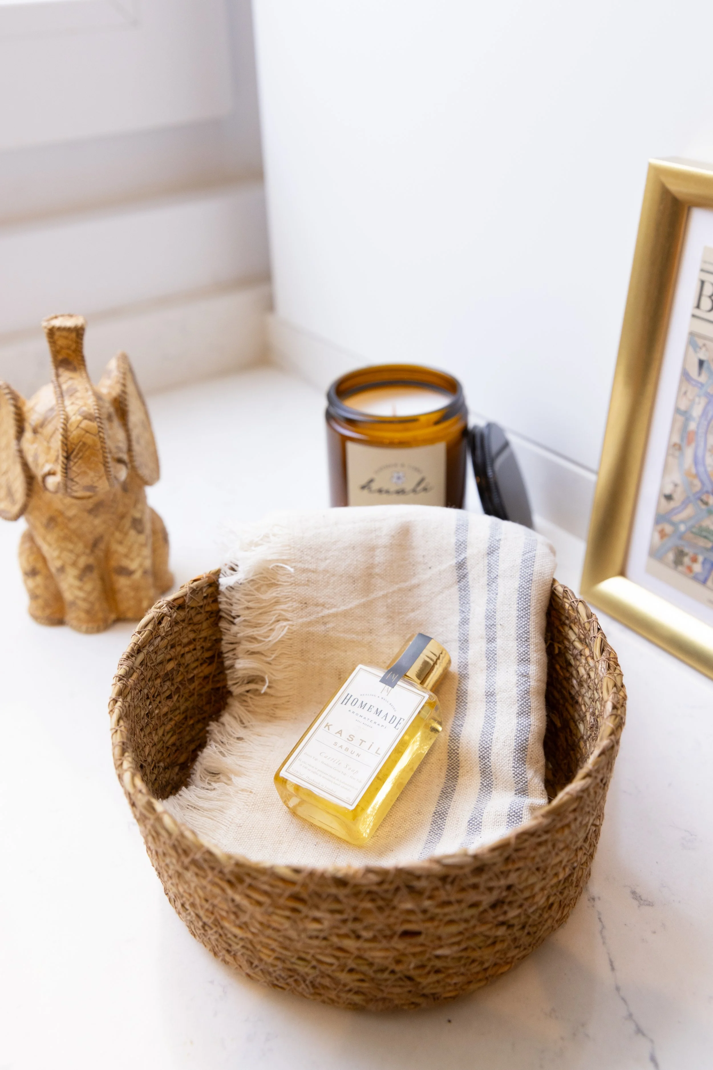 Decorative setup with wicker basket containing a bottle of homemade soap on a cloth, candle, woven elephant, and framed picture on a white surface.