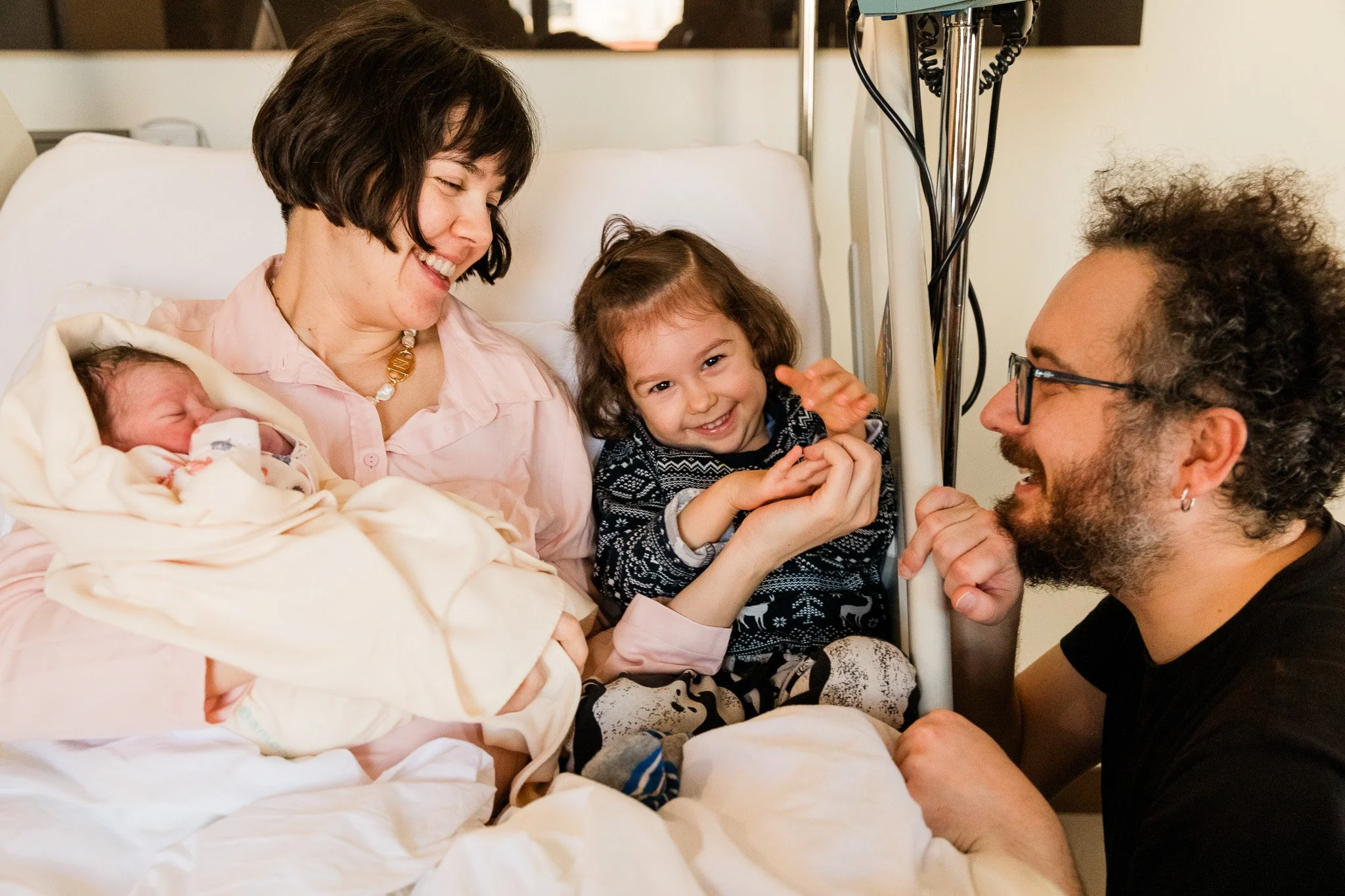 Family in hospital room with newborn baby and child, smiling and interacting.