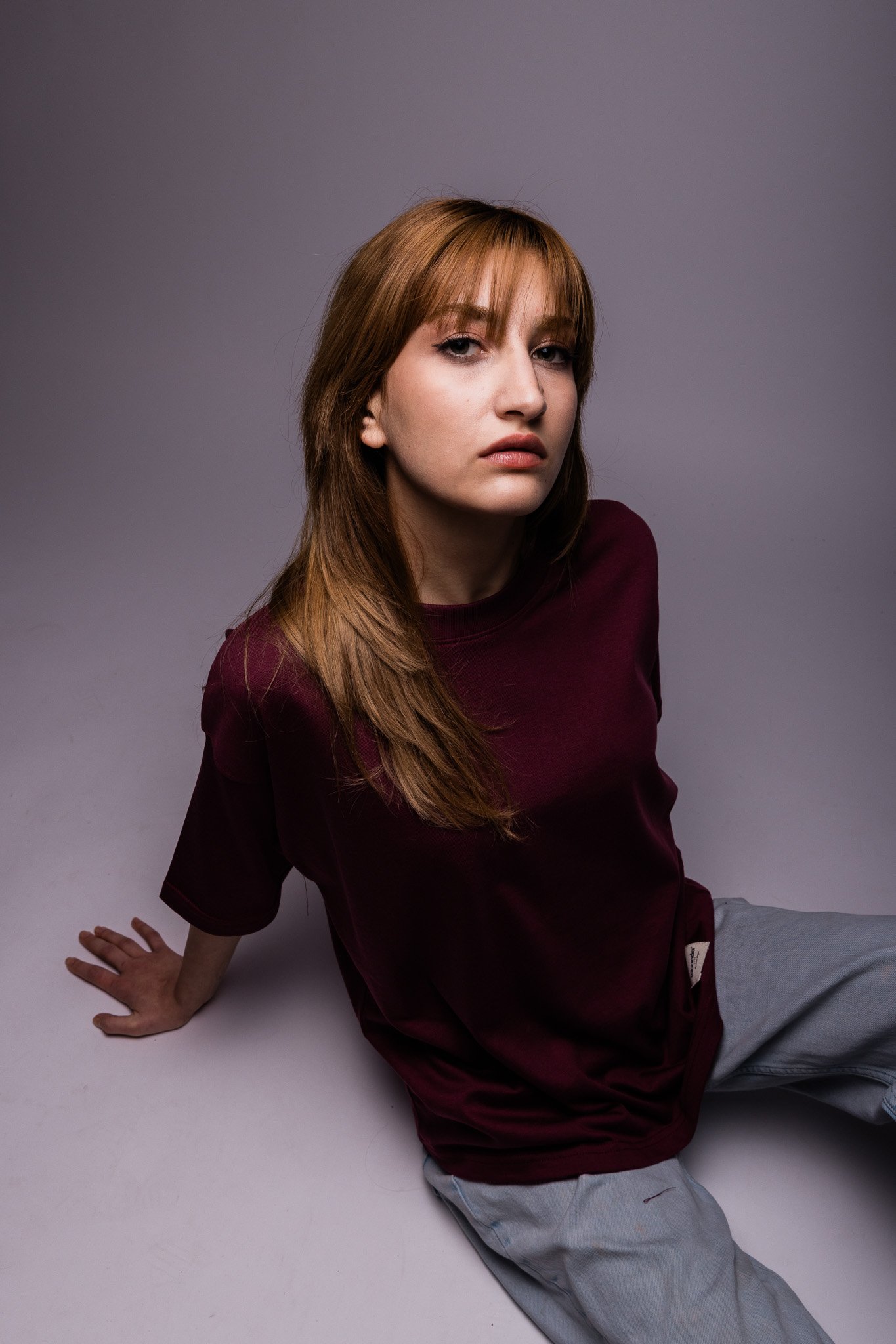 Woman with red hair wearing a burgundy shirt and gray pants sitting on a gray background