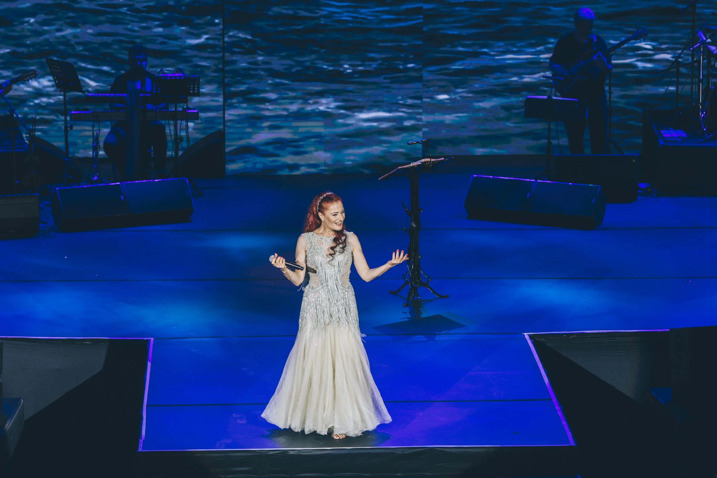Female singer performing on stage with a microphone, wearing a silver gown, with musicians playing instruments in the background and blue stage lighting.
