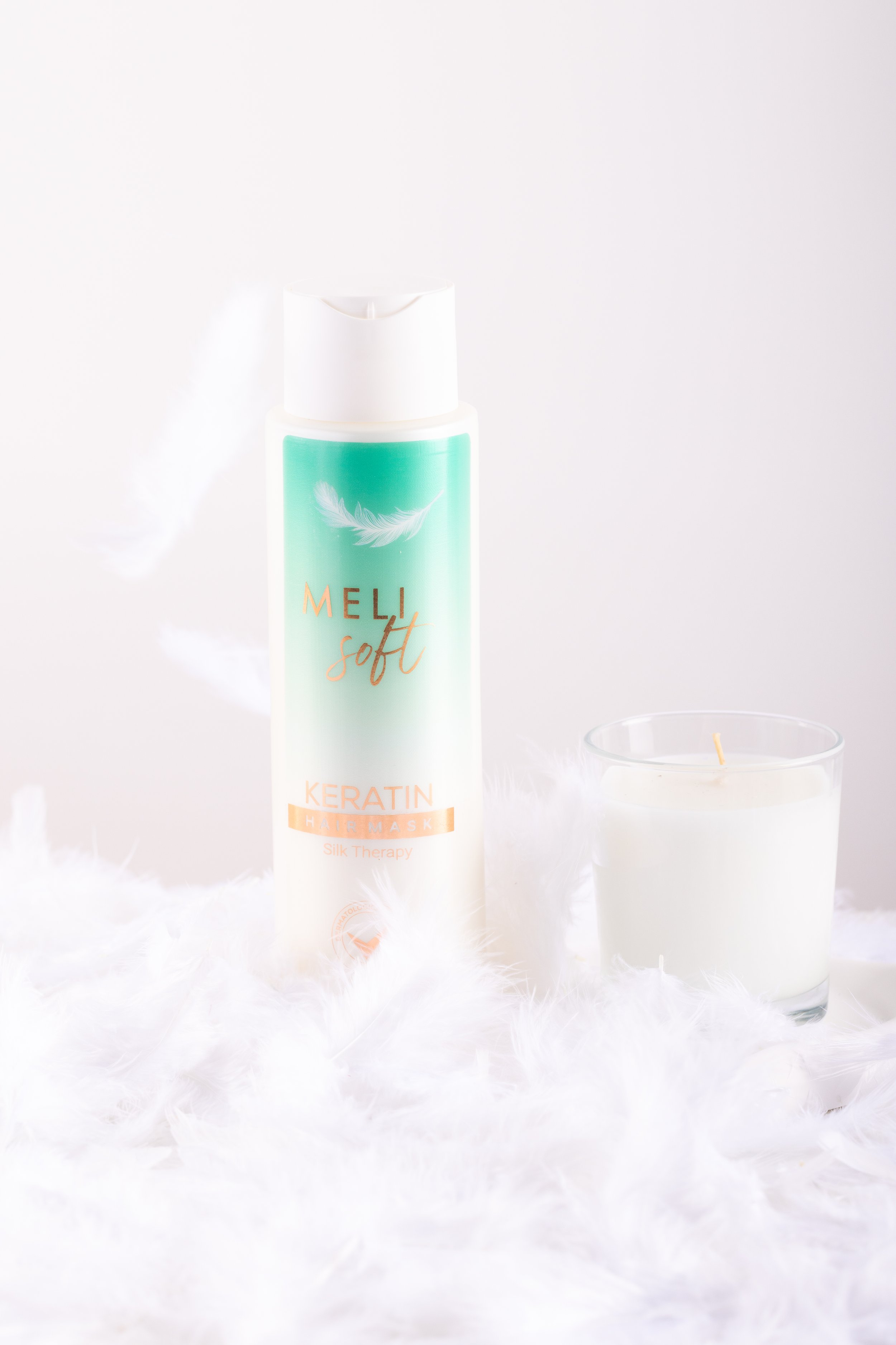 Bottle of Meli Soft Keratin Hair Mask next to a lit white candle on soft feather surface.