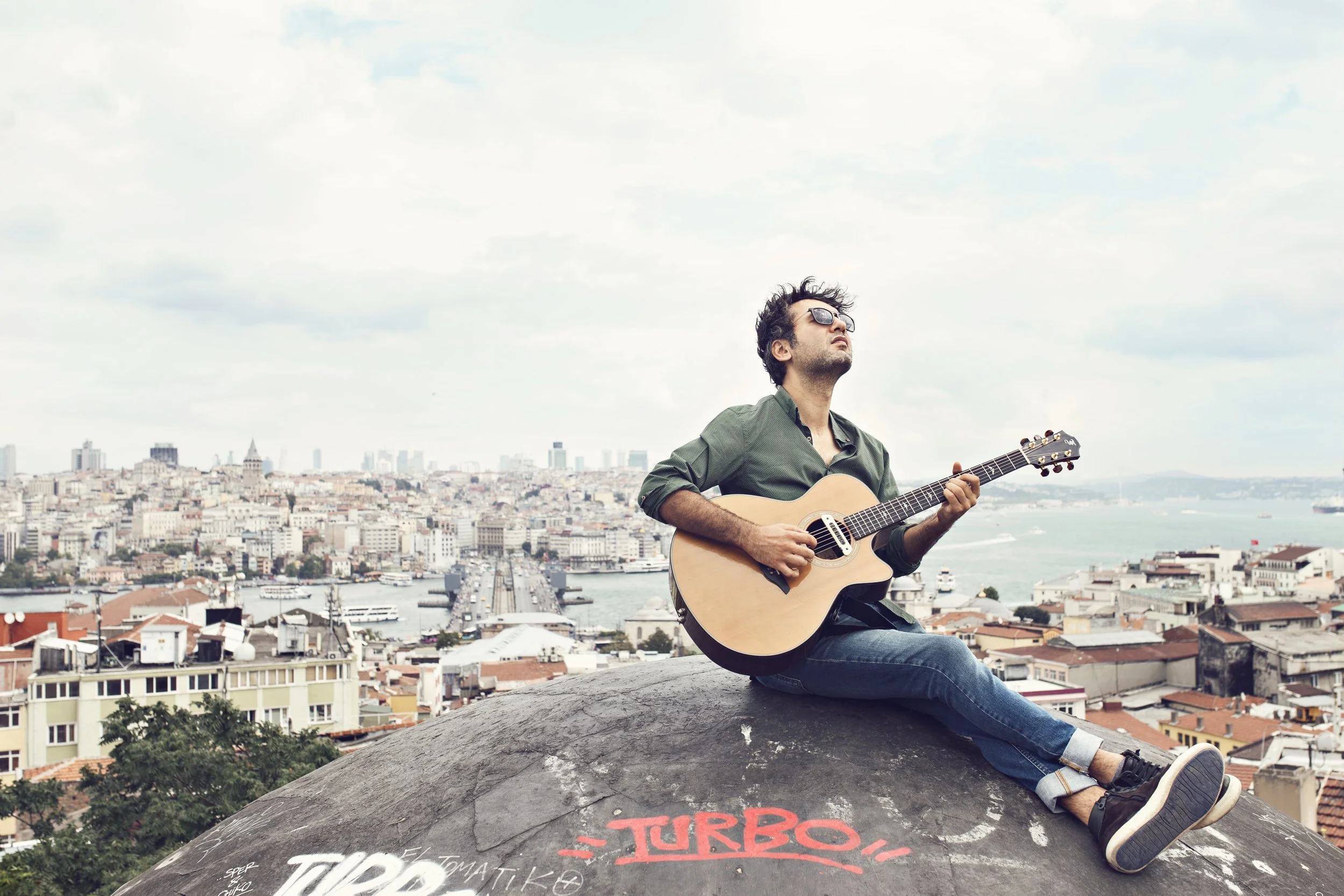 Man sitting on rooftop playing guitar with cityscape background.