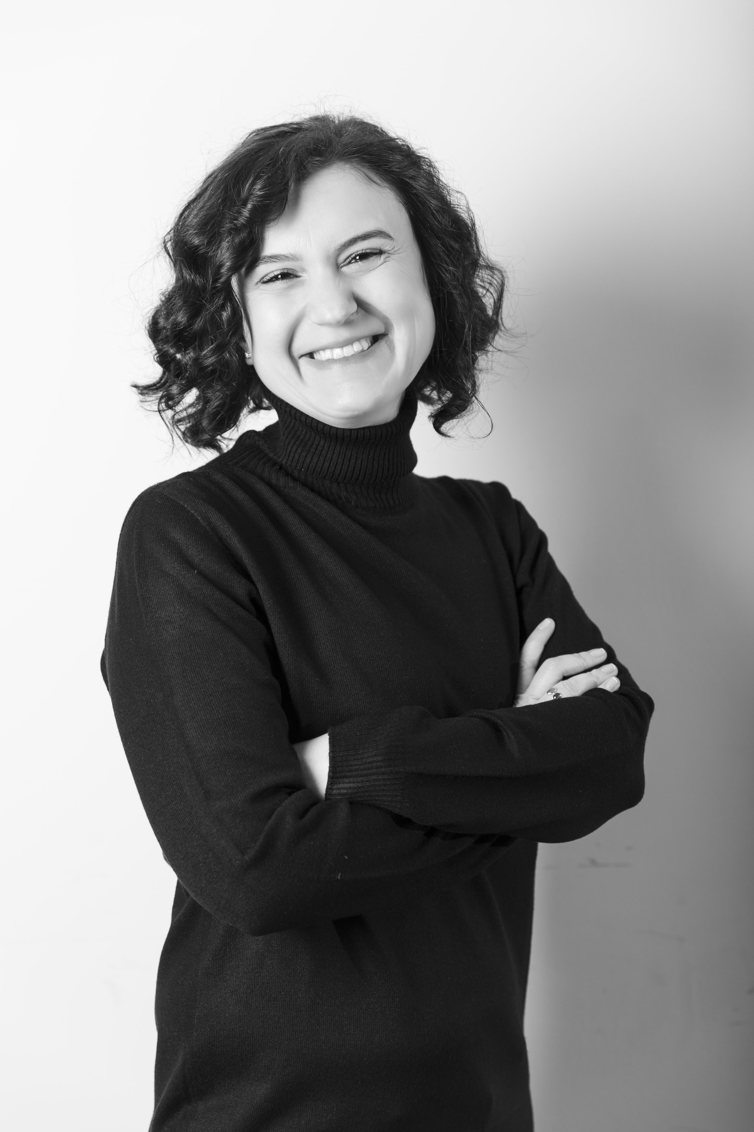 Smiling person with curly hair and a black turtleneck, standing against a plain background.