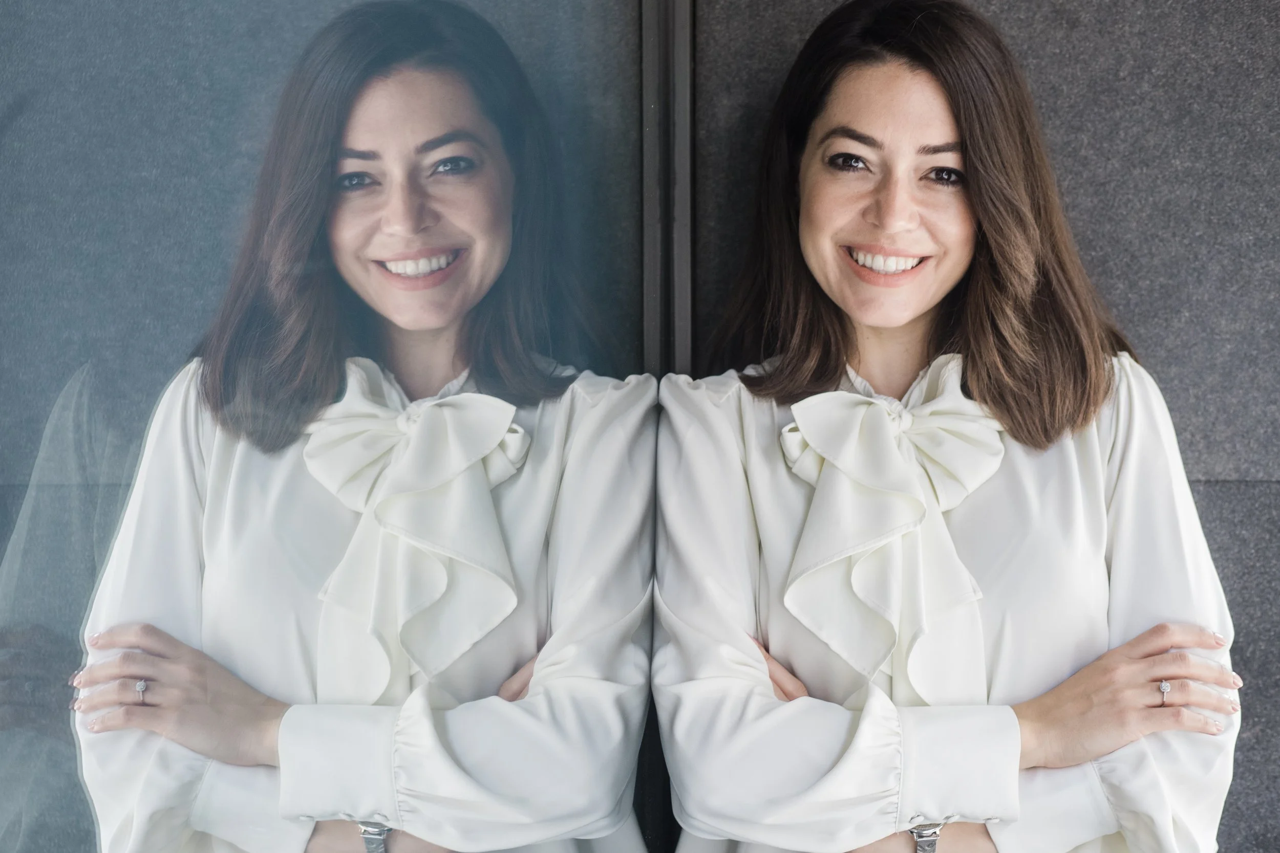 Woman smiling with her reflection in glass, wearing a white blouse with a bow tie, standing against a gray background.