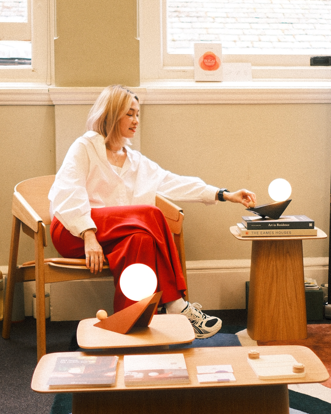 A woman with blonde hair, wearing a white shirt and red pants, sitting in a wooden chair in a room with beige walls and a window. She is reaching out to touch a lamp with a round light on a small table with books and decorative objects.