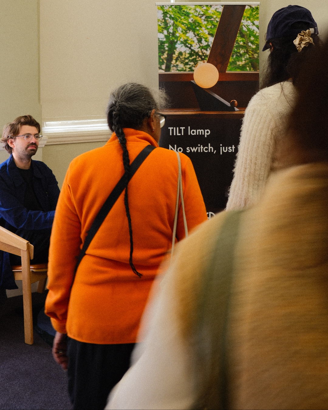 People attending a presentation about a TILT lamp; a woman with gray hair and an orange jacket is facing the poster, and a man with glasses and a dark shirt is sitting in the background.
