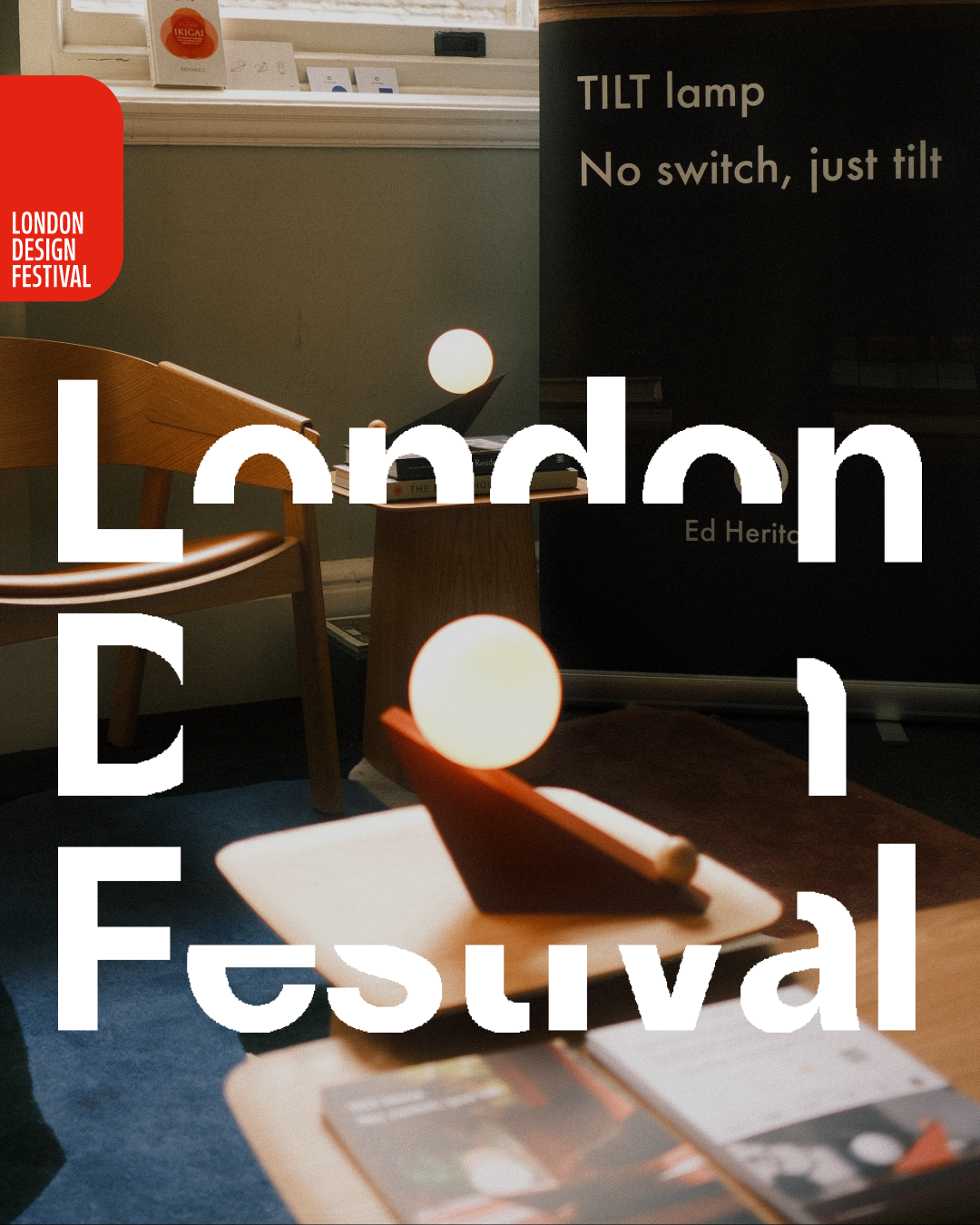Interior setting with wooden chairs, a small round table with books and spherical lamps, and a black poster with white text about a tilt lamp, at the London Design Festival.
