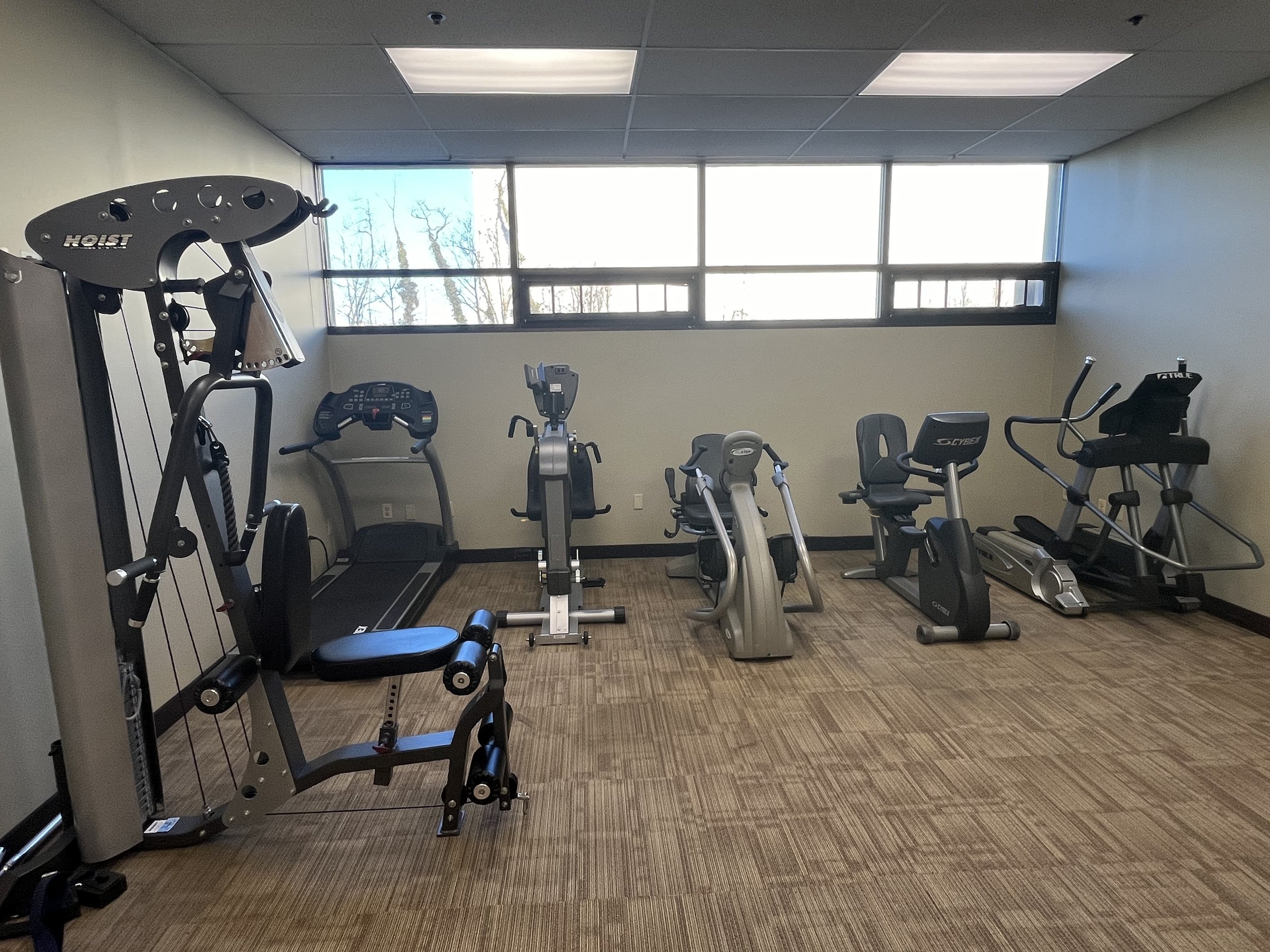 Small gym room with exercise equipment including a multi-station gym, treadmill, stationary bike, elliptical, and recumbent bike under a bright window.