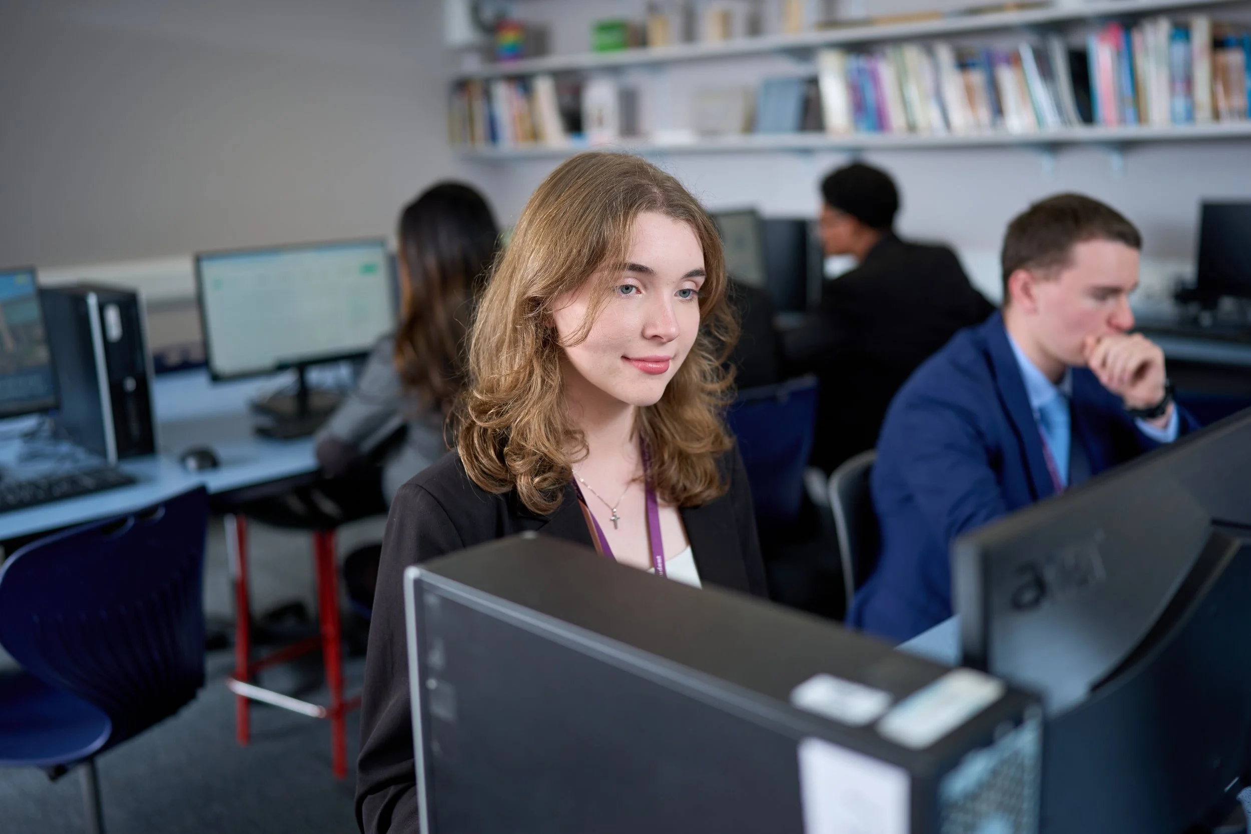 Students working at computers in a classroom, with a smiling woman in the foreground and a man in a blue shirt behind her