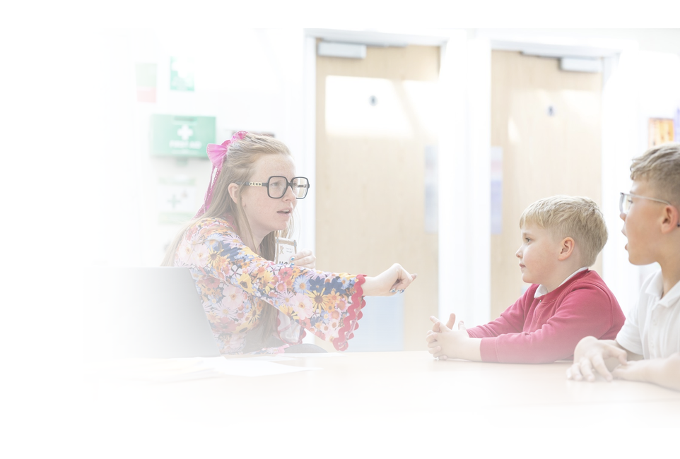 Teacher wearing glasses and a floral blouse speaks with two schoolboys at a classroom table.