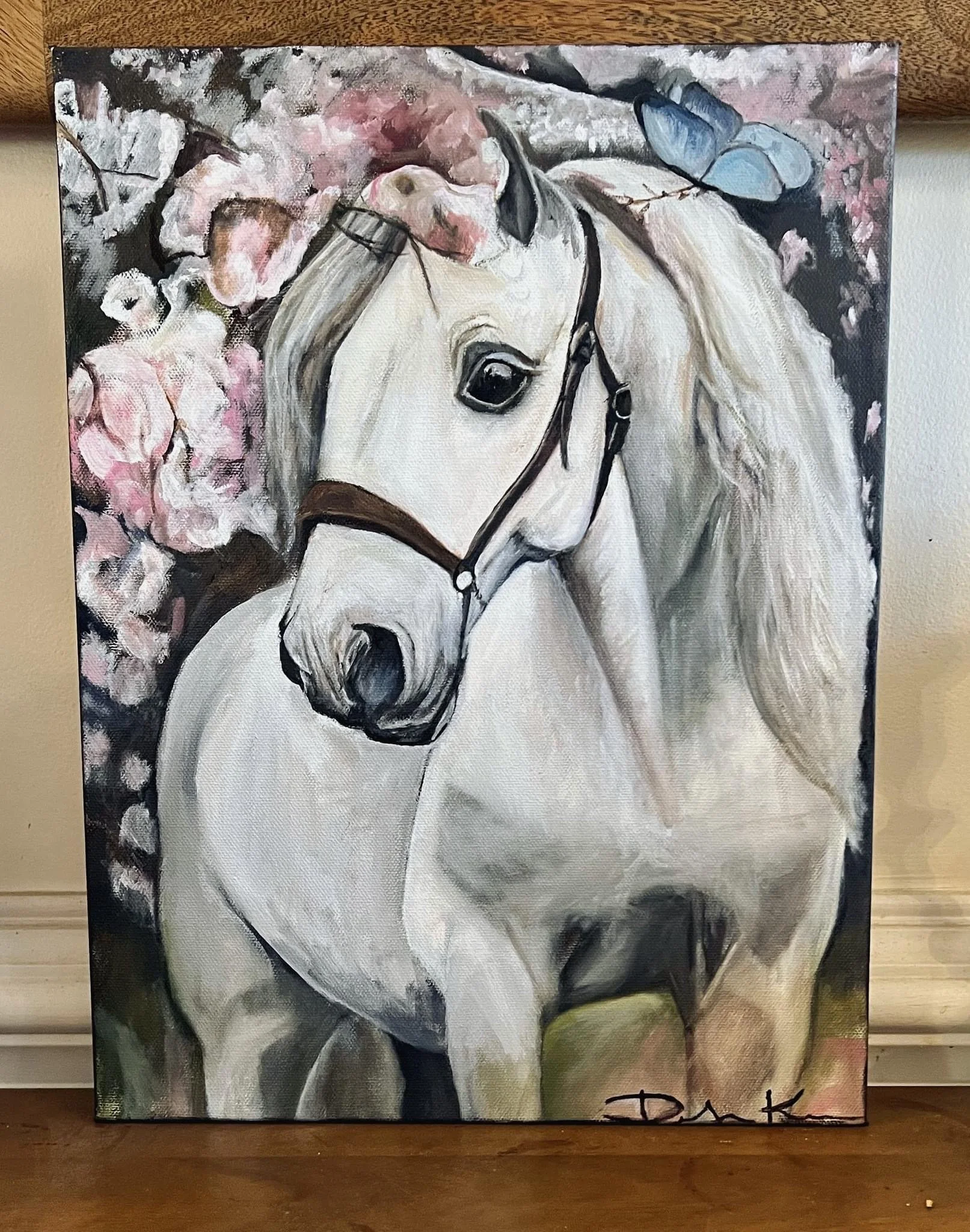 Painting of a white horse with a flowing mane, black bridle, and large expressive eye, surrounded by pink and white blossoms and a butterfly in the background.